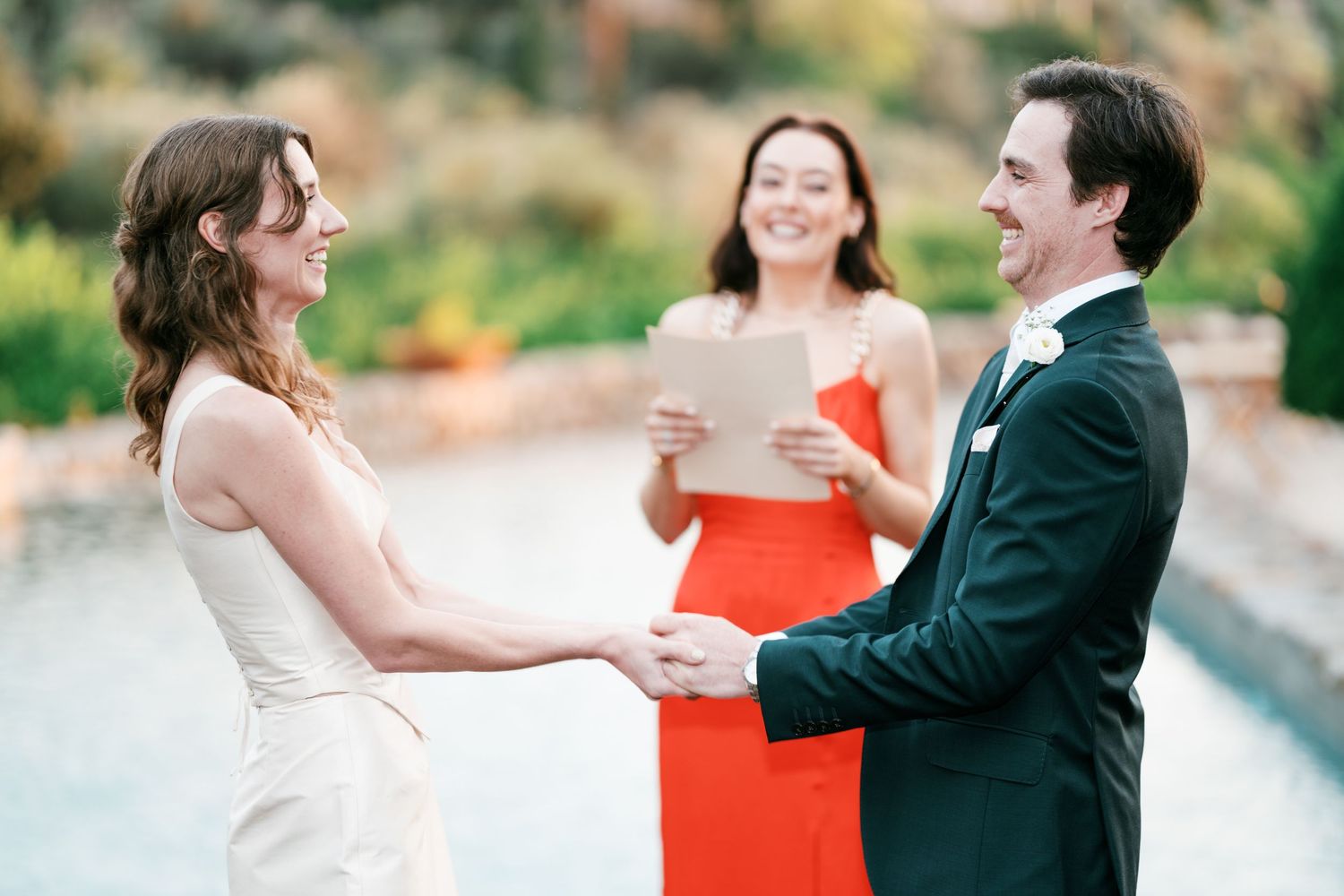 Bride in a white dress and groom in a dark suit hold hands during an outdoor wedding ceremony as the officiant in red smiles and reads.