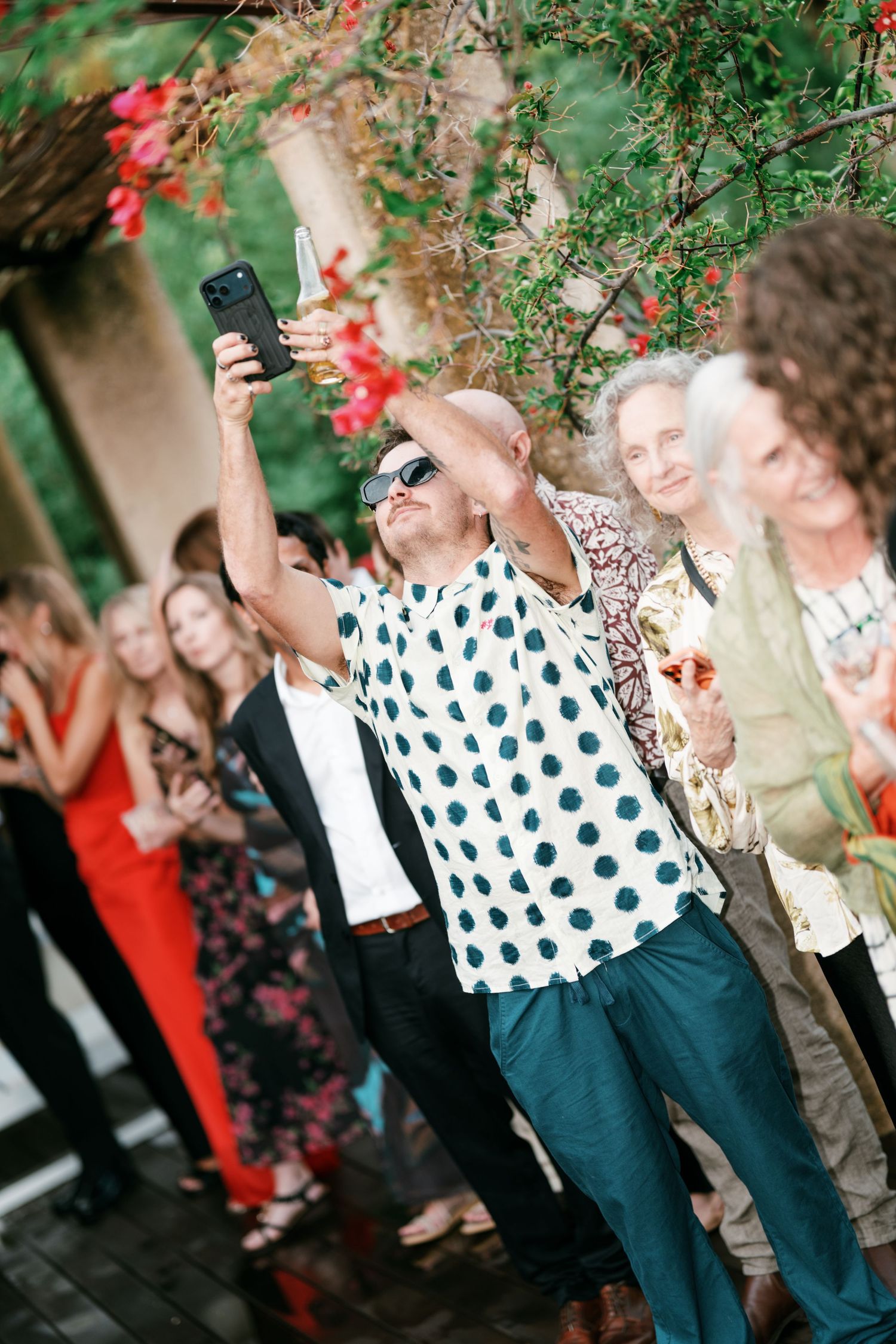 Man in a polka dot shirt and sunglasses raises a phone and glass to celebrate among smiling wedding guests under blooming flowers.