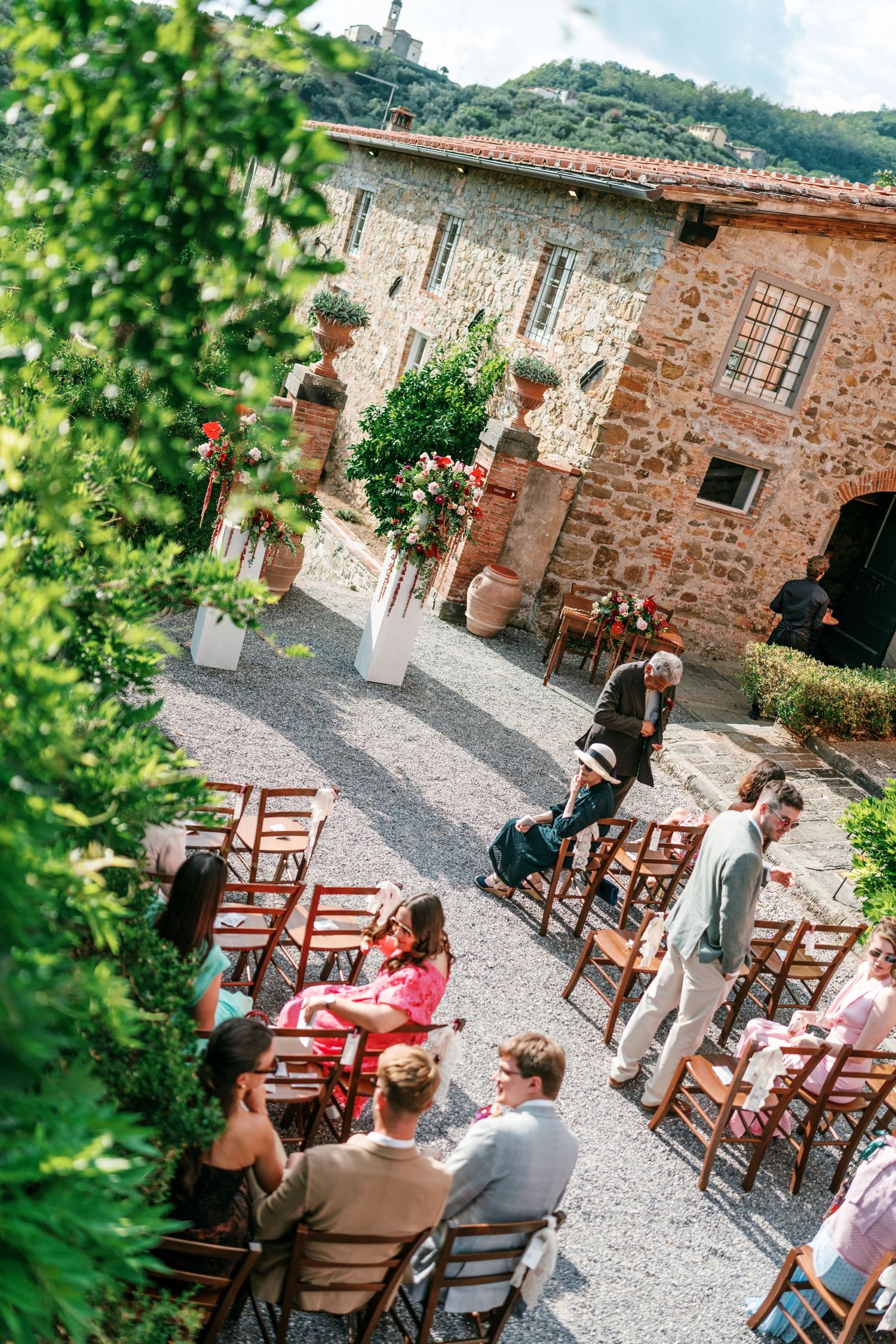Wedding guests seated on wooden chairs along a gravel aisle outside a rustic stone villa, with flower arrangements and greenery in Tuscany.