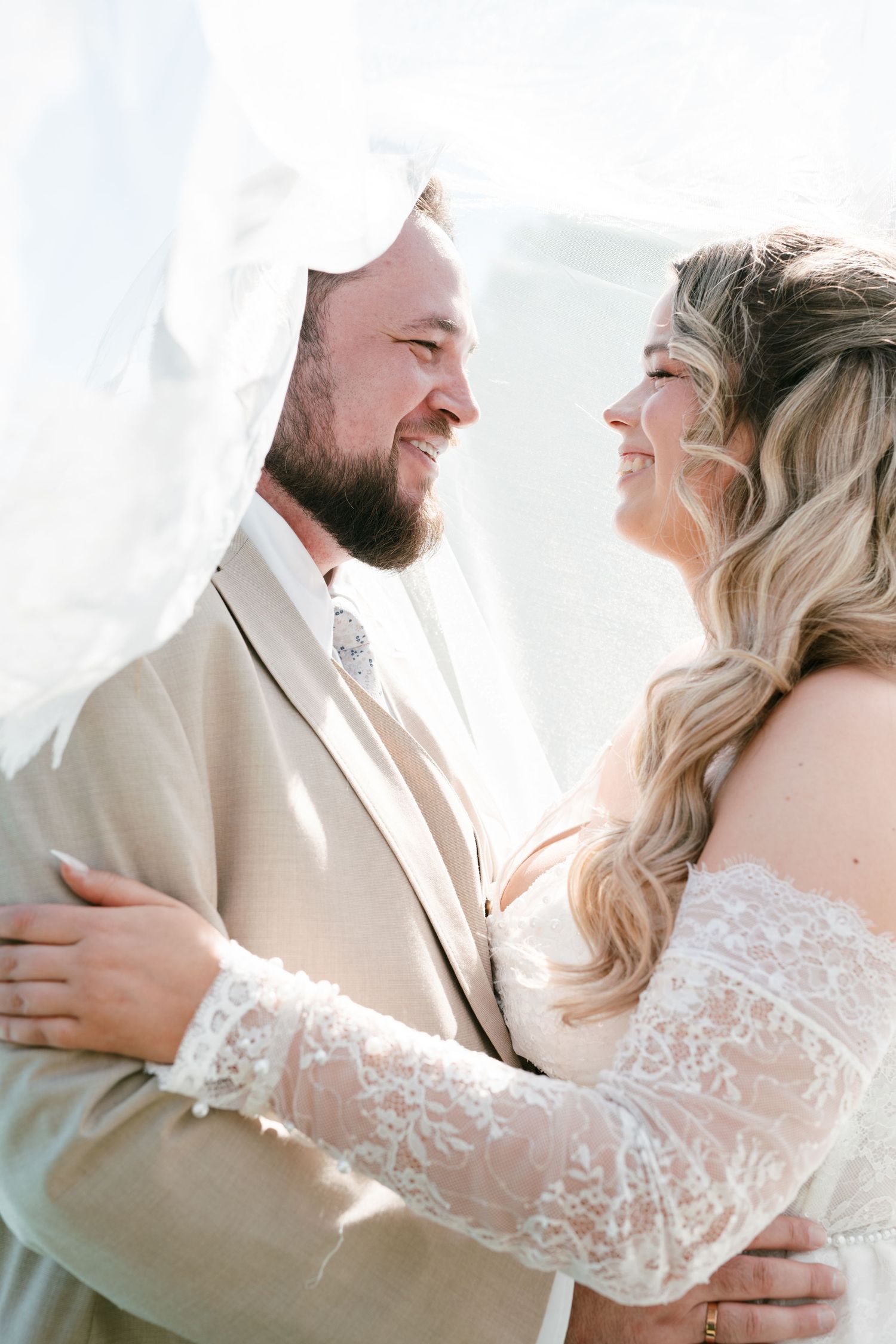 Bride and groom smiling at each other under a veil, groom in tan suit, bride in lace off-shoulder gown in Mallorca.