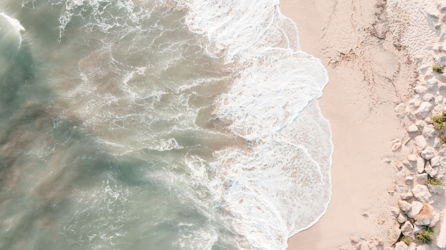Aerial view of ocean waves gently washing onto a sandy beach with white foam and scattered rocks along the shoreline. Mallorca wedding film