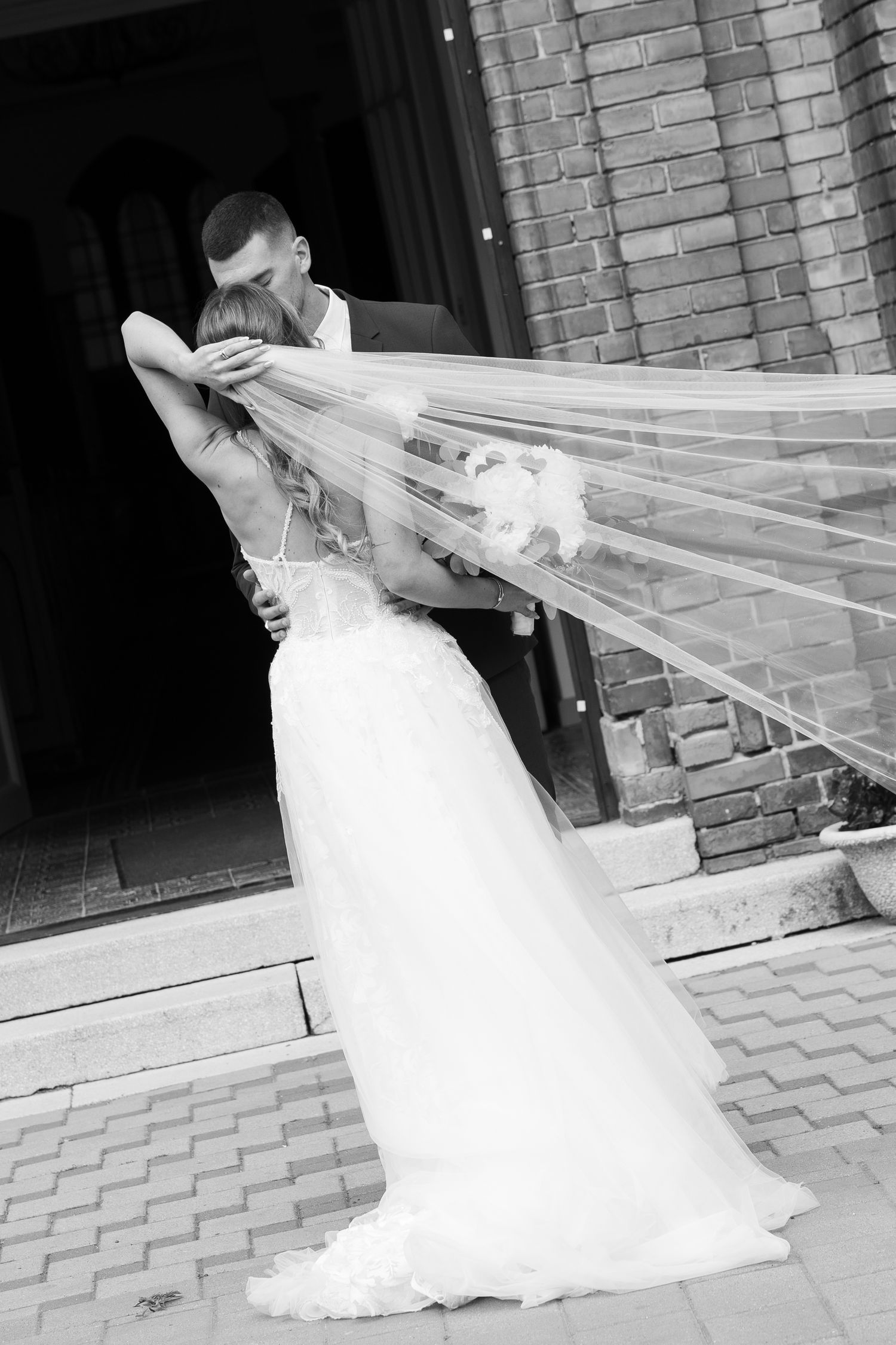 Black and white photo of bride and groom kissing at church entrance with veil flowing in wind. Mallorca wedding photographer and filmmaker.