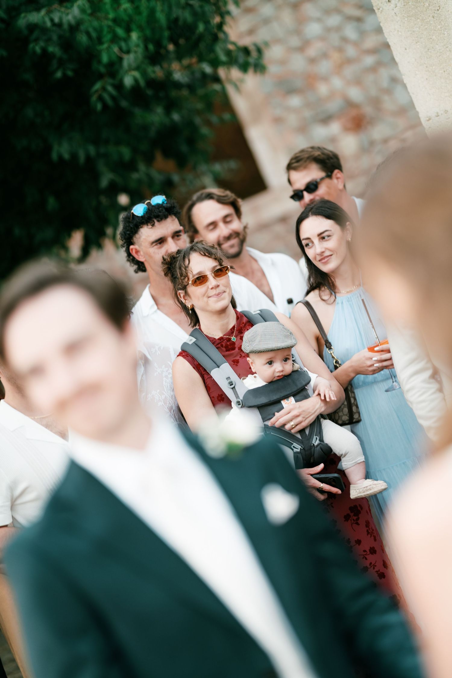 Wedding guests smile and watch a ceremony outdoors, including a woman carrying a baby in a carrier, with a blurred groom in a green suit in 