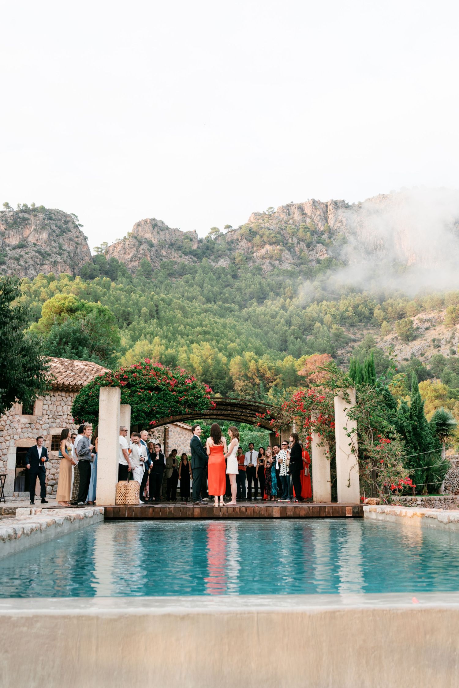 Outdoor wedding ceremony by a stone-edged pool beneath a rustic pergola, with guests gathered and misty green mountains rising behind.