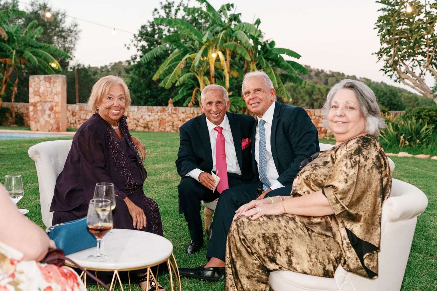 Four smiling older adults in elegant attire sit on white lounge chairs around a small table with wine glasses at an outdoor garden party.