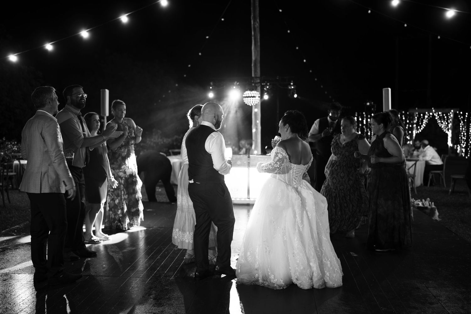 Black and white photo of a bride in a ball gown and groom dancing at a wedding reception while guests watch under string lights.