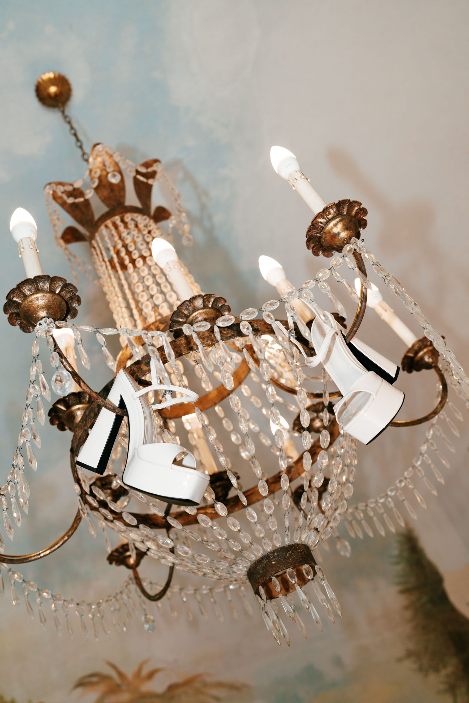 White platform sandals hanging from a vintage crystal chandelier with candle-style bulbs against a painted blue ceiling mural.