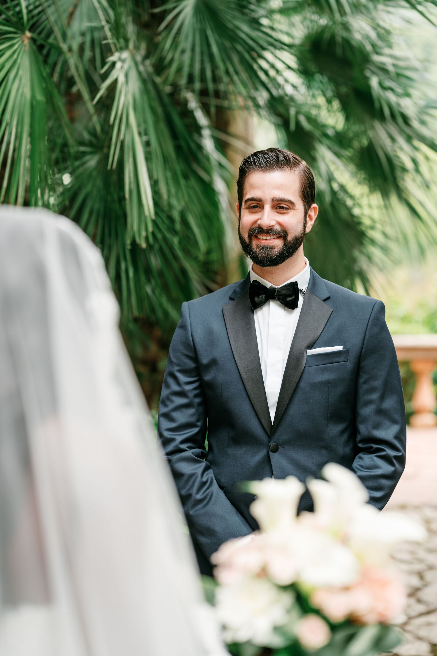 Smiling bearded groom in a navy tuxedo and bow tie stands among palm trees as the veiled bride and a bouquet appear in the foreground.