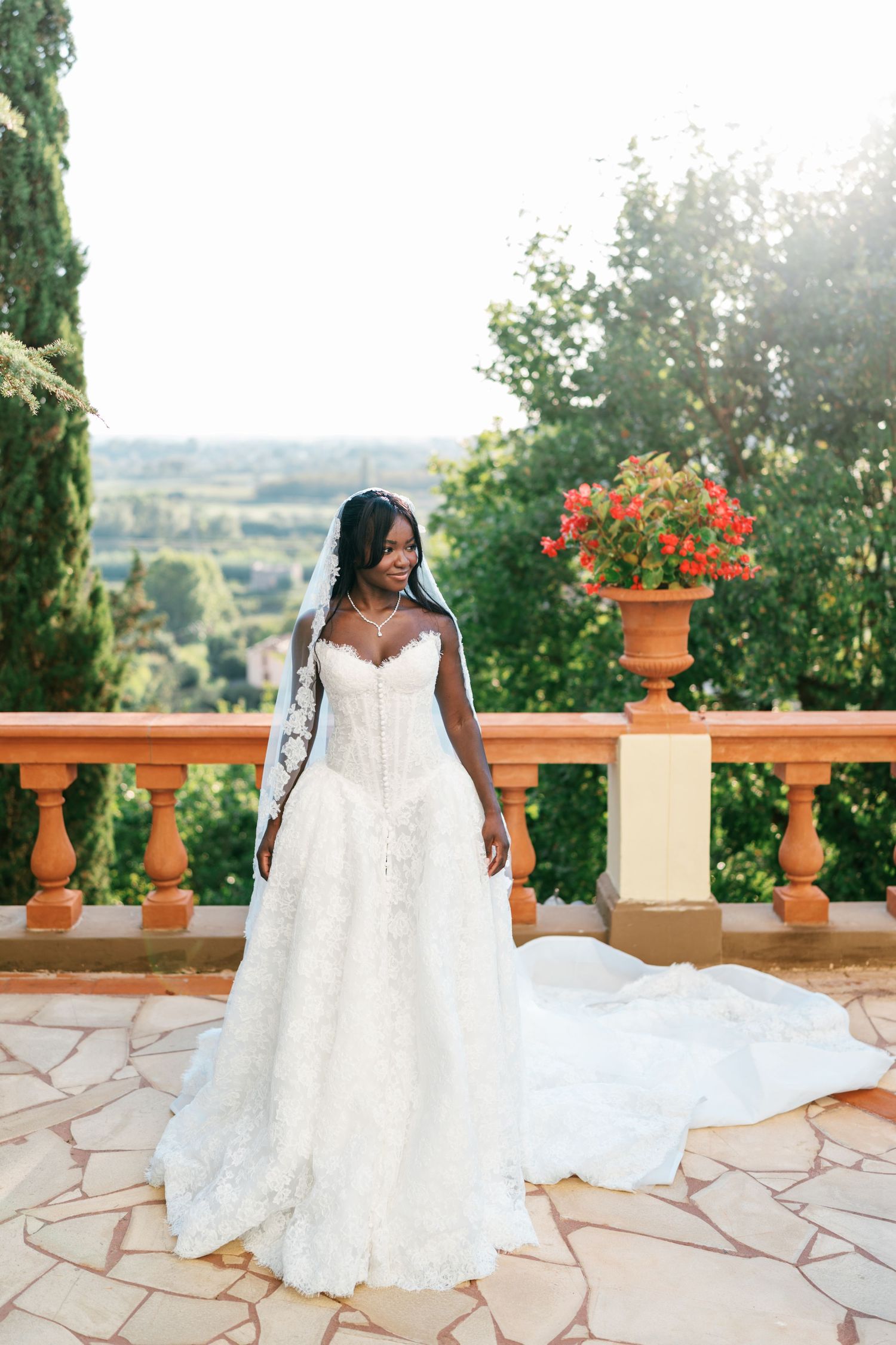 Bride in a white lace strapless gown and long veil stands on a stone terrace with a terracotta balustrade overlooking a scenic countryside.