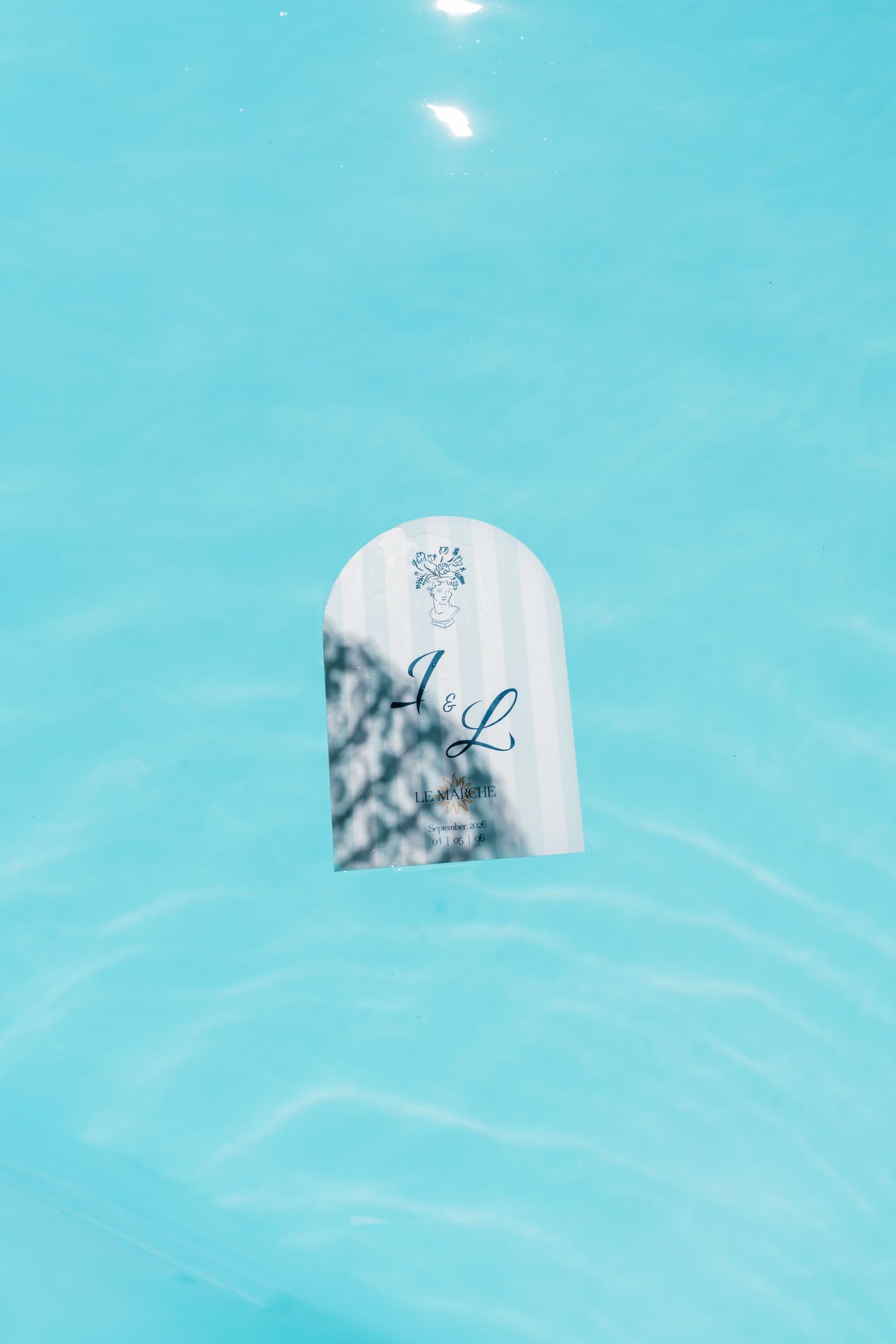 Elegant wedding invitation with A&L monogram floating in a bright turquoise blue swimming pool.