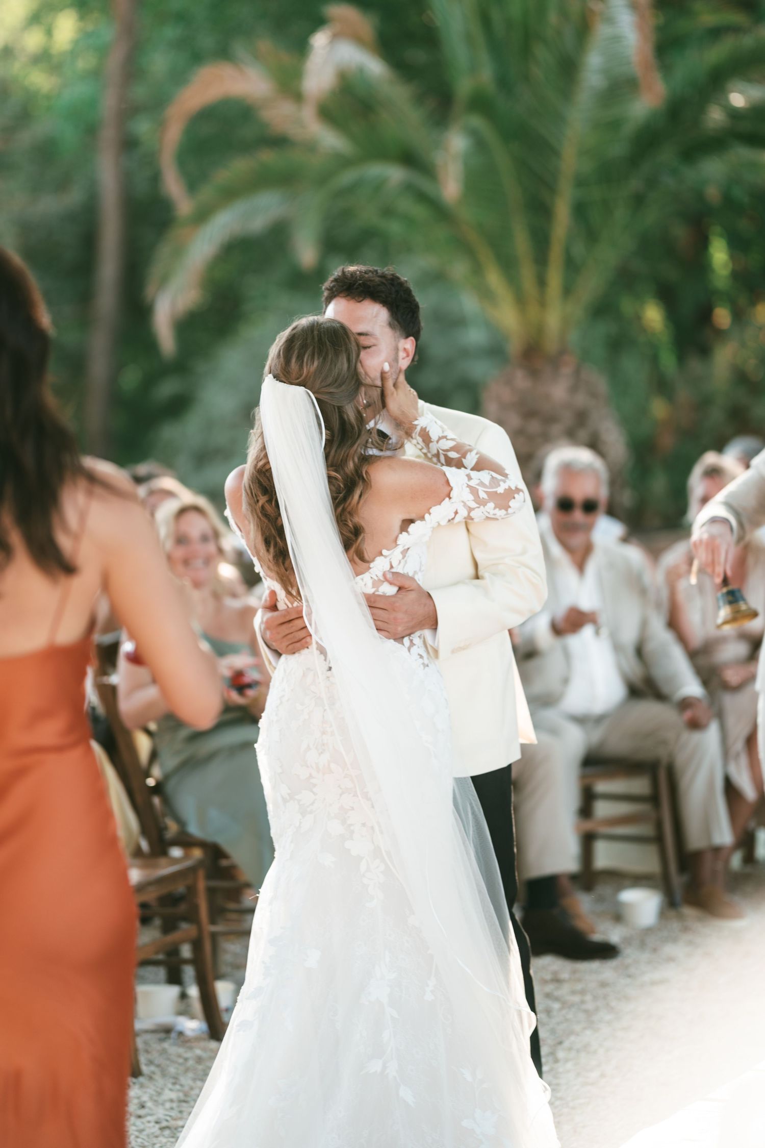 Bride and groom share romantic first dance kiss outdoors with smiling wedding guests seated in background at sunset.