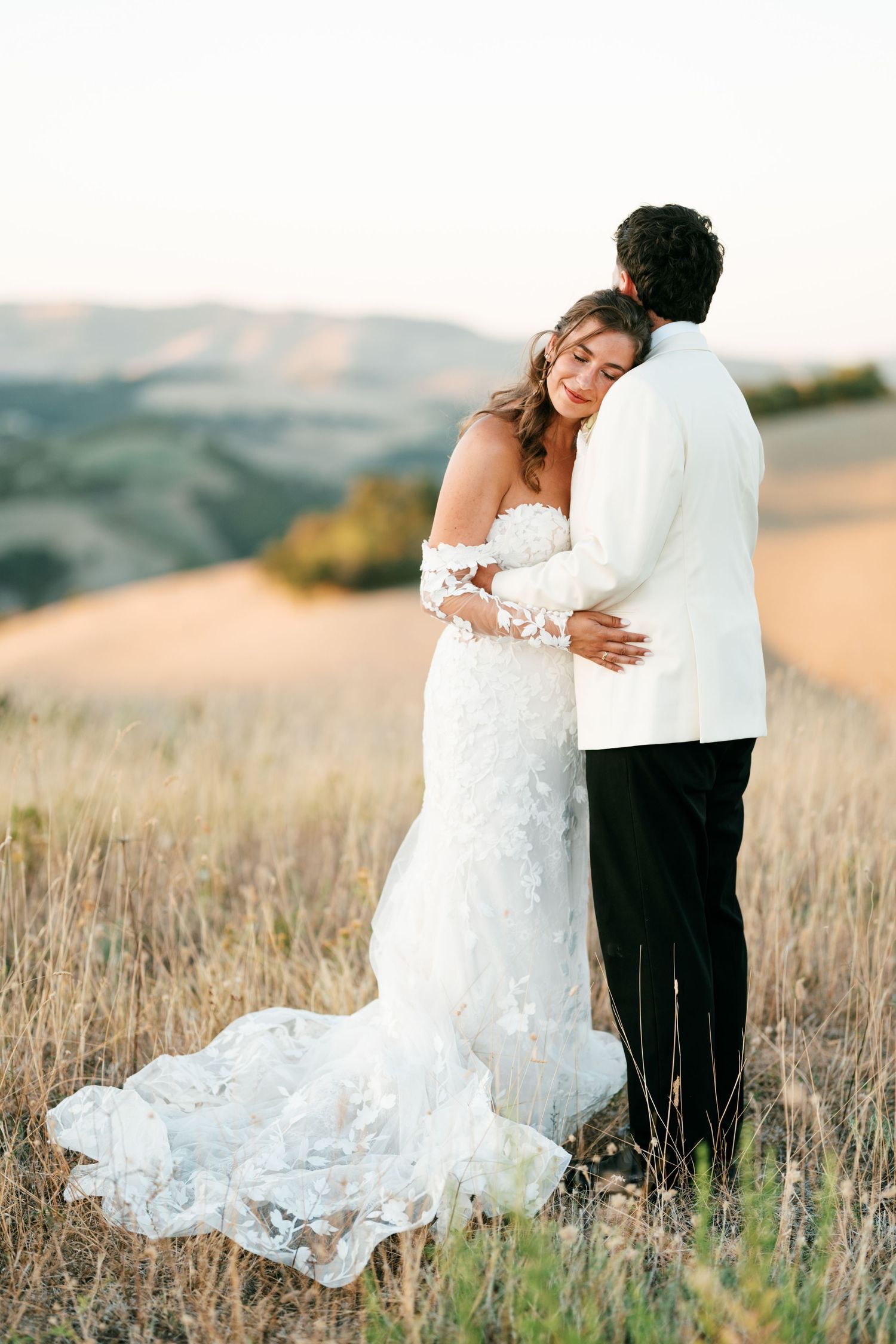 Bride in floral lace gown embraces groom in white jacket at golden hour in rolling hills.