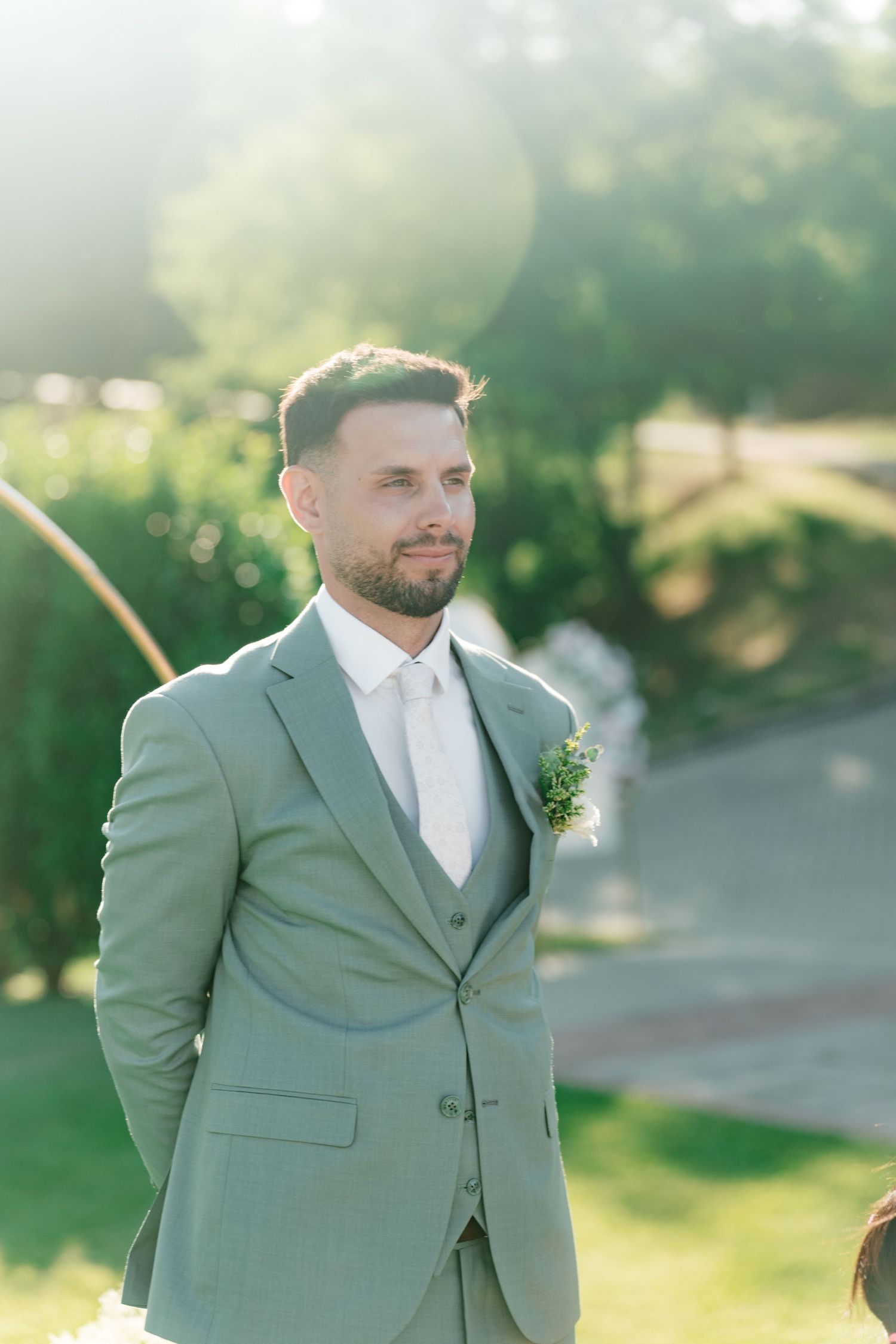 Handsome groom in a sage green suit with boutonniere standing outdoors in golden sunlight. Italy wedding photographer and filmmaker