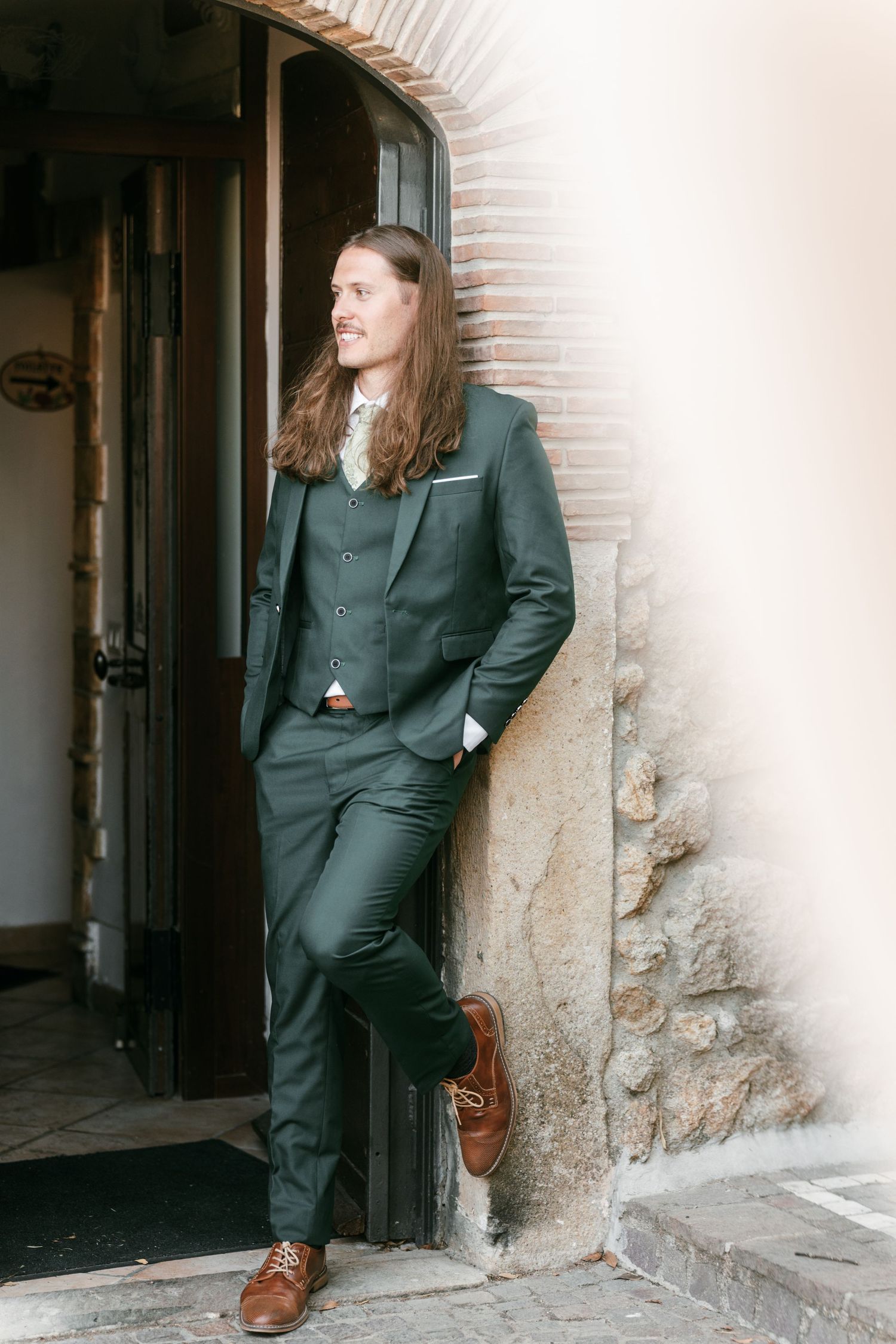 Stylish groom with long hair in a dark green three-piece suit leaning against a rustic stone doorway. Tuscany wedding photographer, filmmake