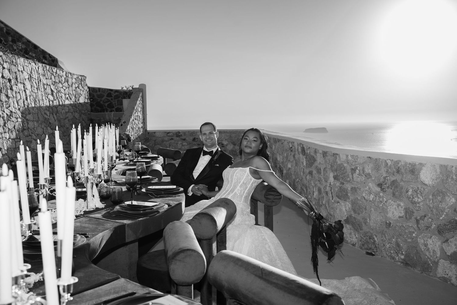 Black and white wedding photo of bride and groom seated at elegant candlelit table with ocean view.