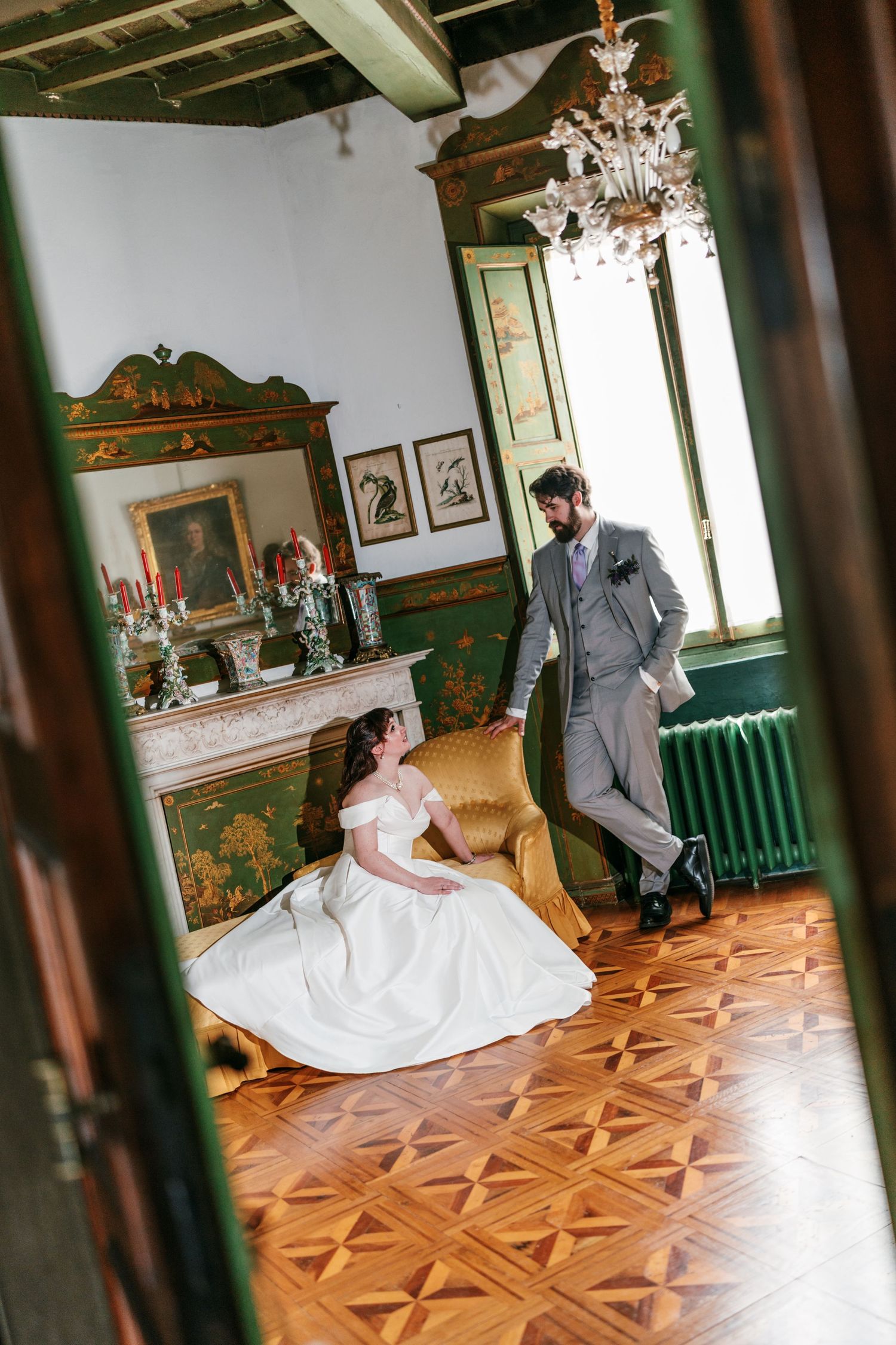Bride in an off-shoulder white gown sits on a gold armchair while the groom in a gray suit stands in an ornate room with a chandelier.