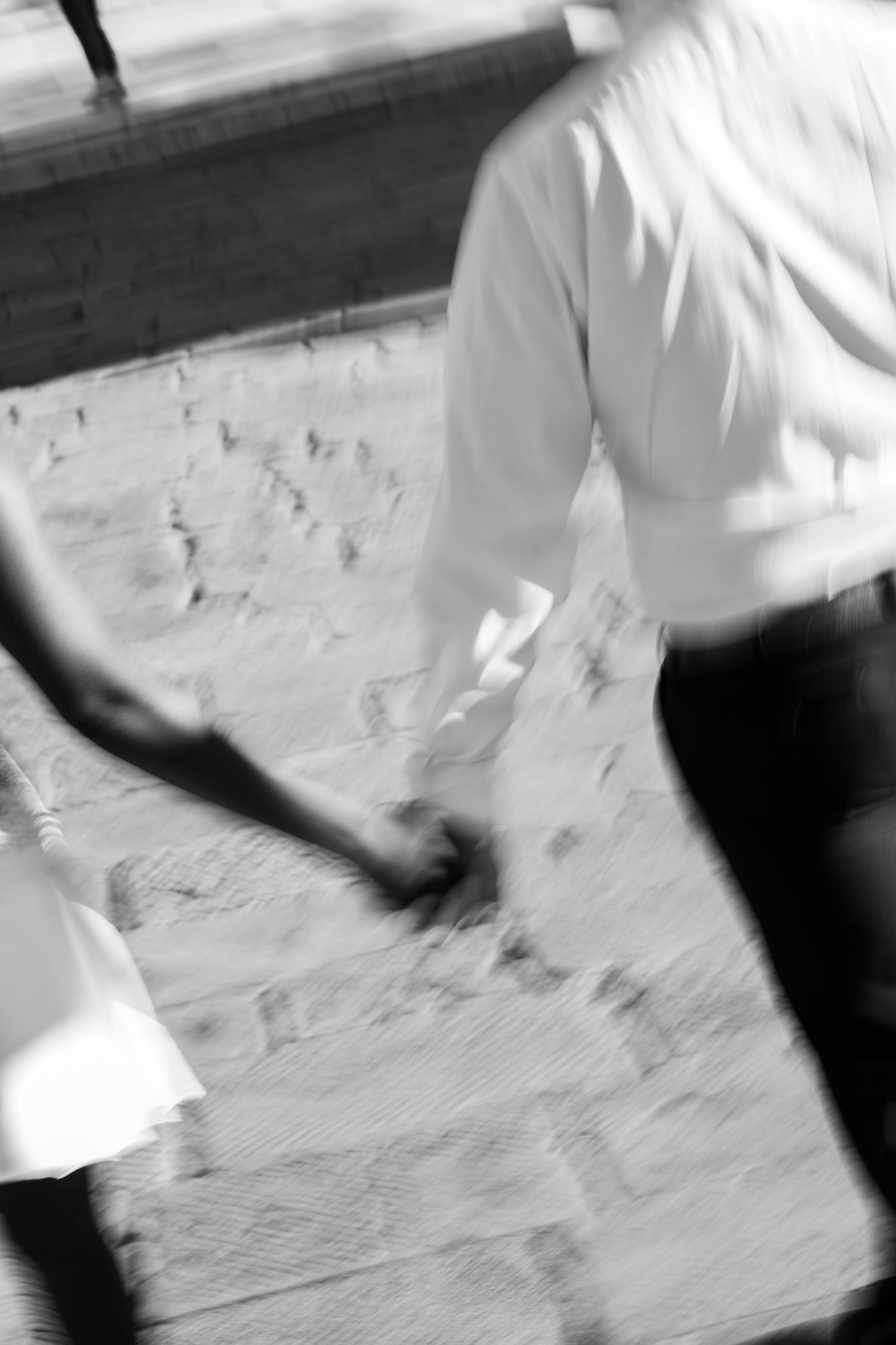Black and white photo of a couple holding hands while walking along a sunlit stone pavement in motion blur.