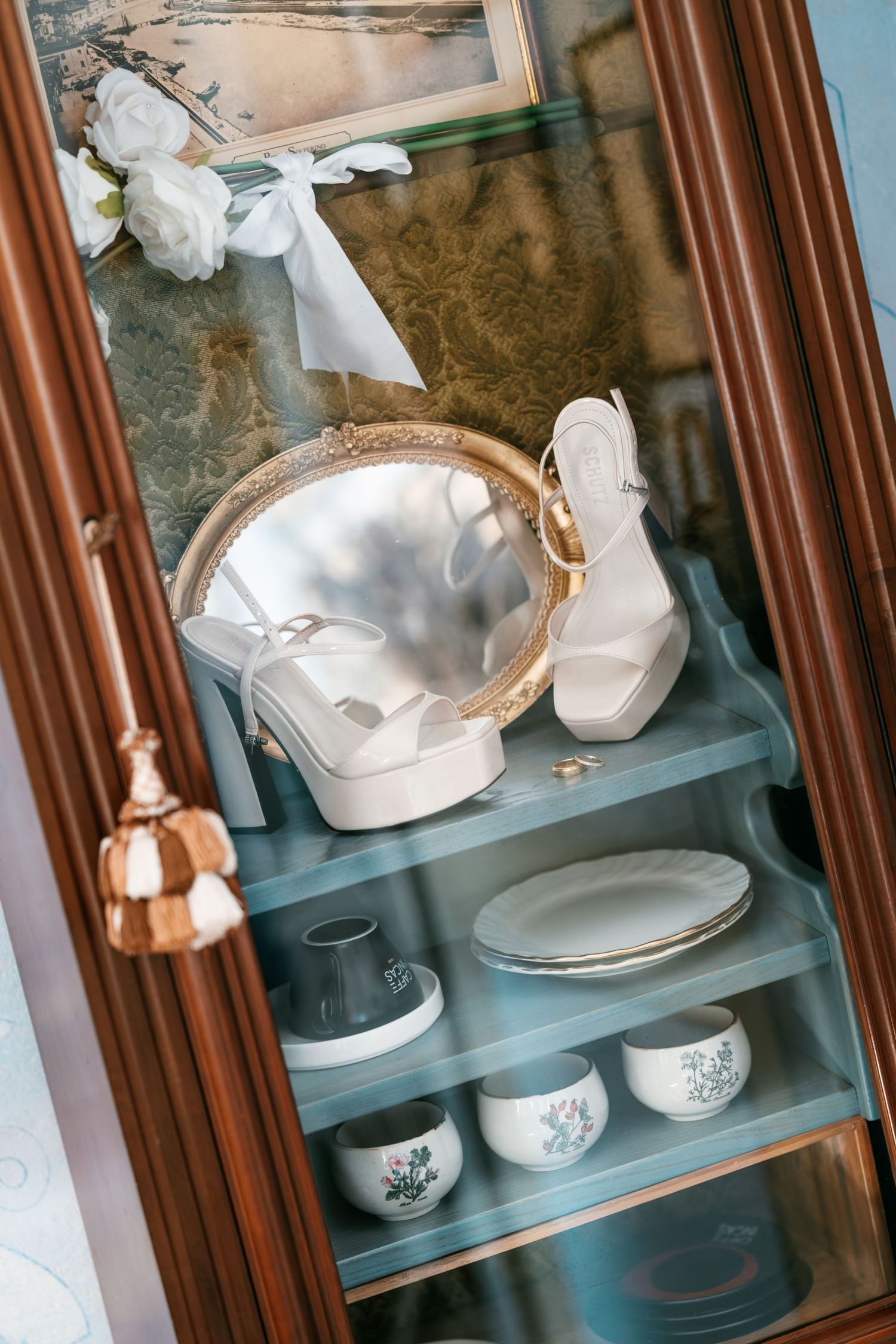 White bridal heels and a round gold mirror displayed in a wooden china cabinet with a white floral bow and dishes.