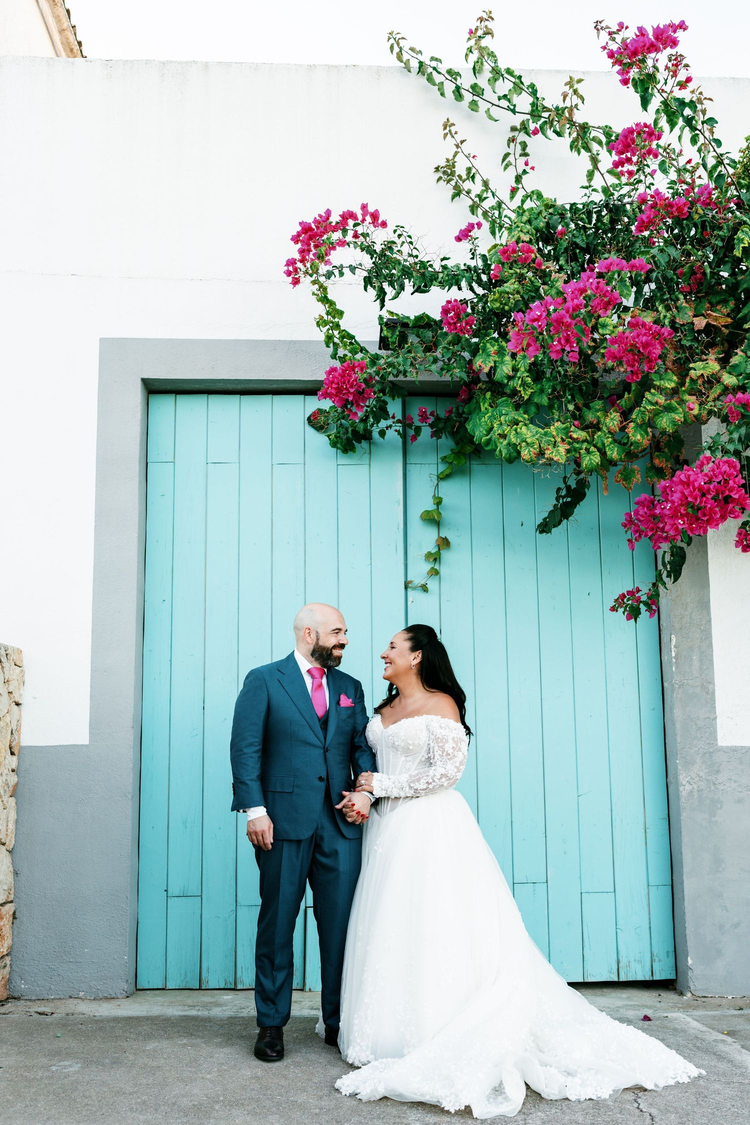 Bride in a white off-the-shoulder lace gown and groom in a blue suit hold hands before a turquoise wooden door with pink bougainvillea.