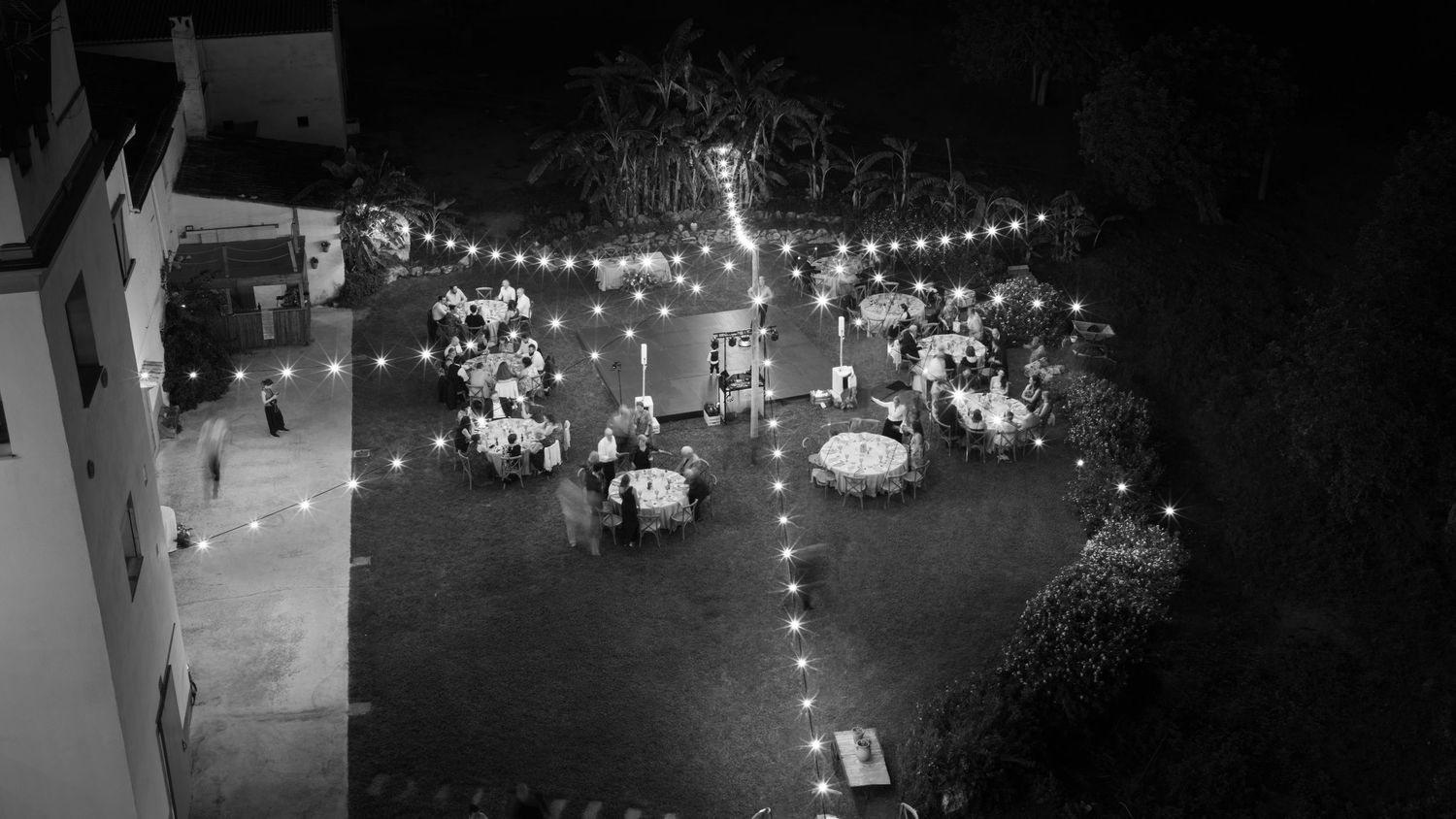 Black and white aerial view of an outdoor evening reception with guests at round tables beneath glowing string lights on a lawn.
