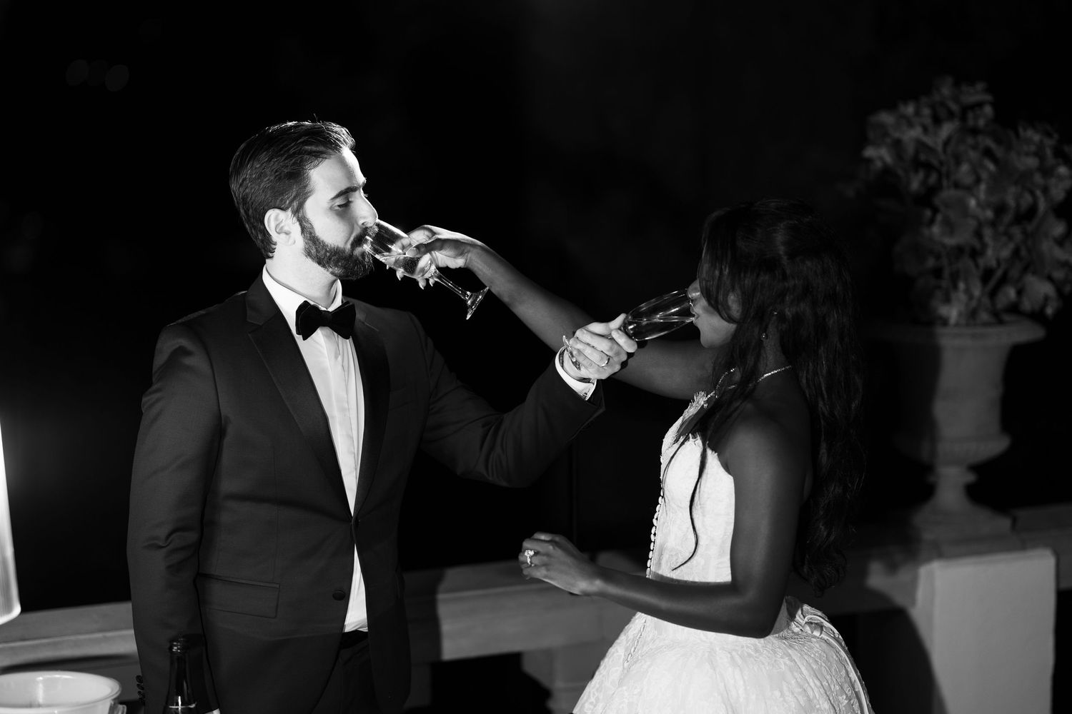 Black and white photo of a bride and groom linking arms as they toast with champagne glasses during their wedding celebration.