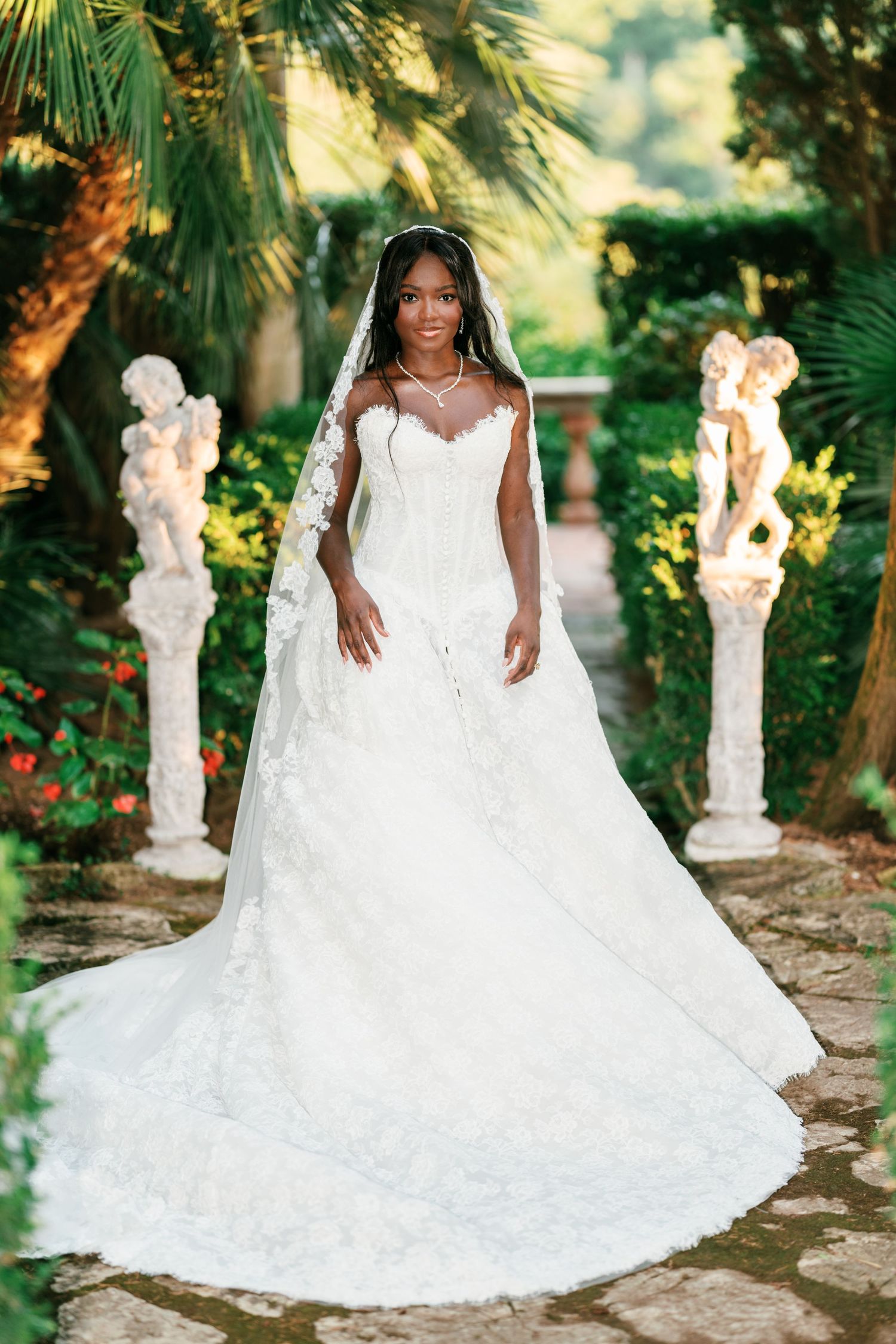 Bride in a lace wedding gown and long veil poses on a stone garden path beside statues and lush greenery.