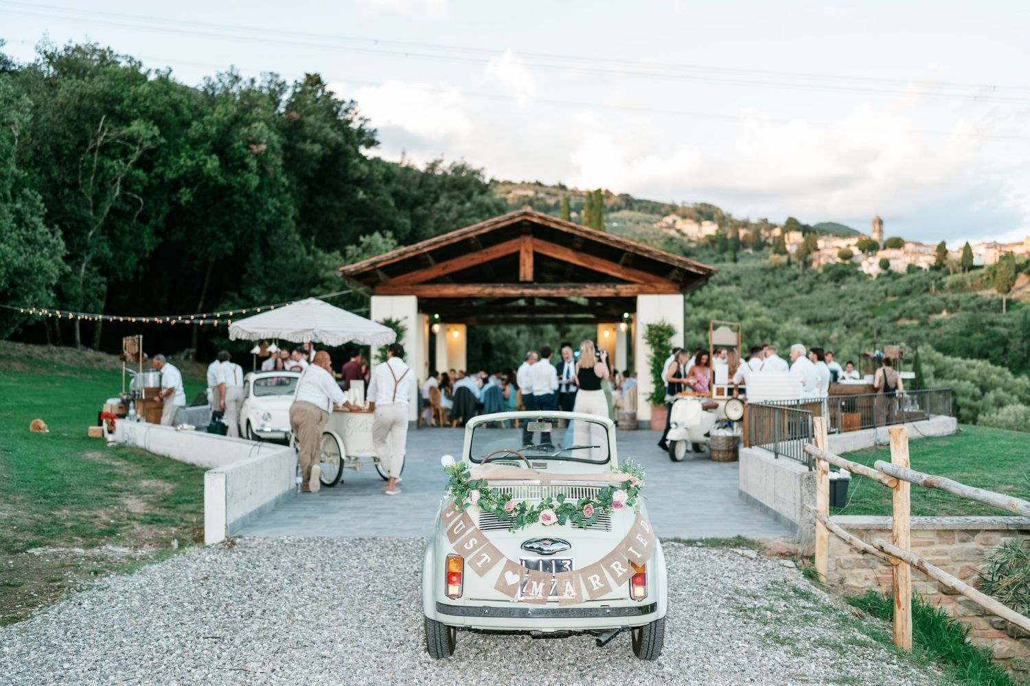 Vintage white Fiat 500 decorated with greenery parked at a rustic Tuscan wedding venue with guests gathered beneath a wooden pavilion.