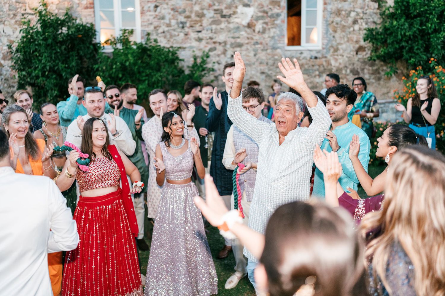Wedding guests in colorful Indian attire clap and cheer outdoors in front of a stone building during a lively celebration.