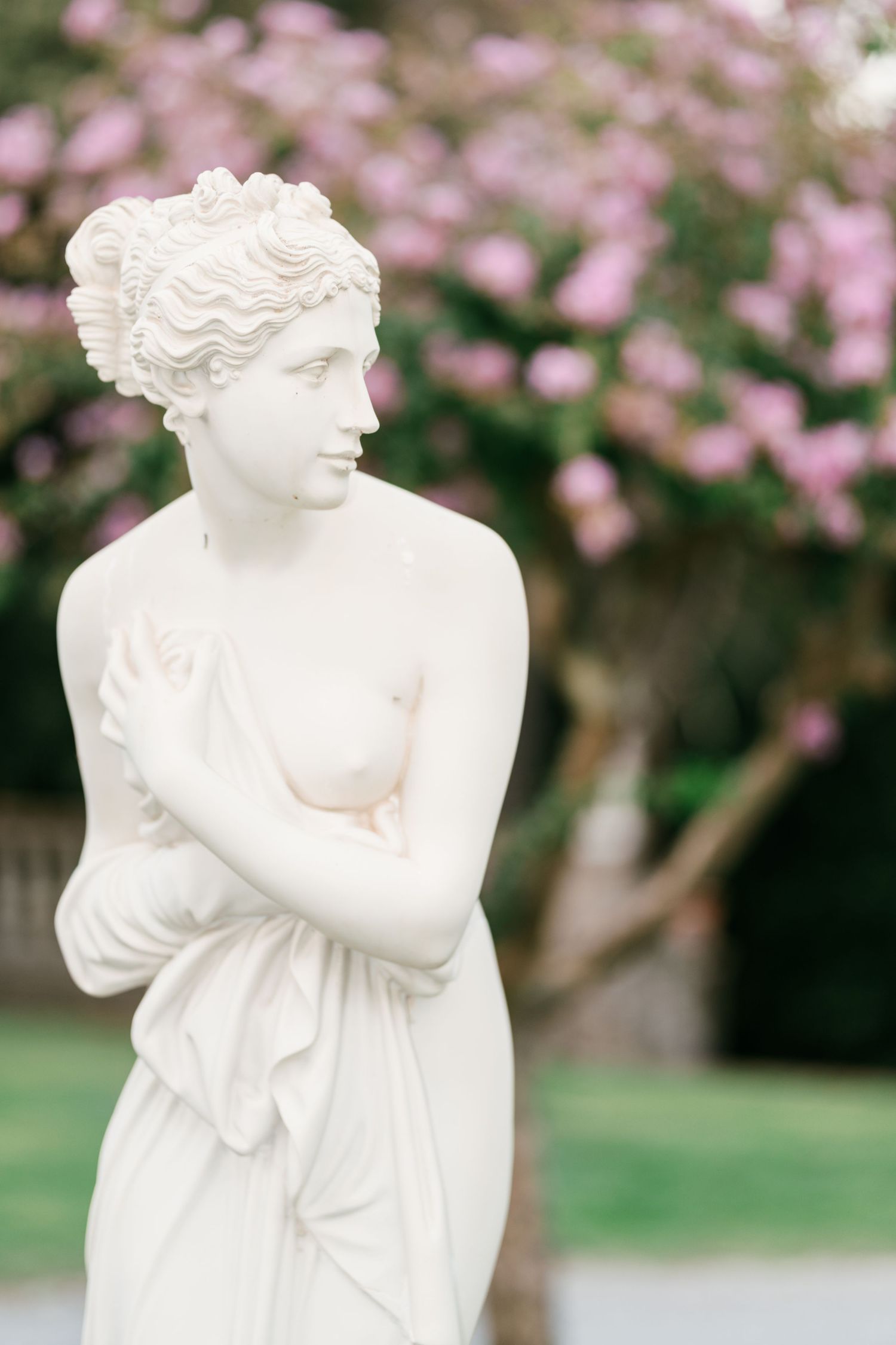 White marble statue of a classical woman with an elegant updo, draped in cloth, in a garden with pink blossoms behind.