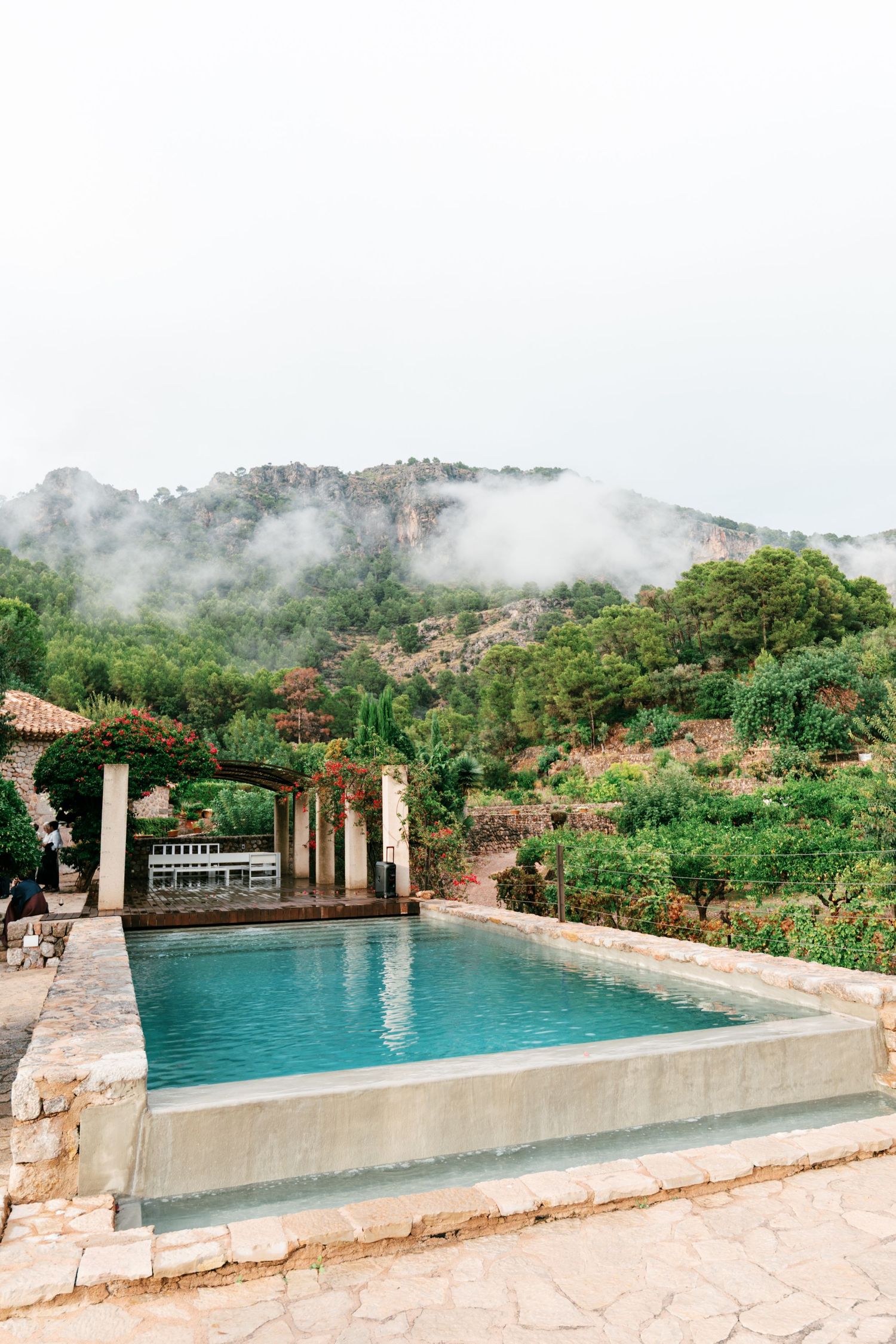 A stone-edged swimming pool with turquoise water sits beside a pergola, overlooking misty green mountains and lush Mediterranean gardens.