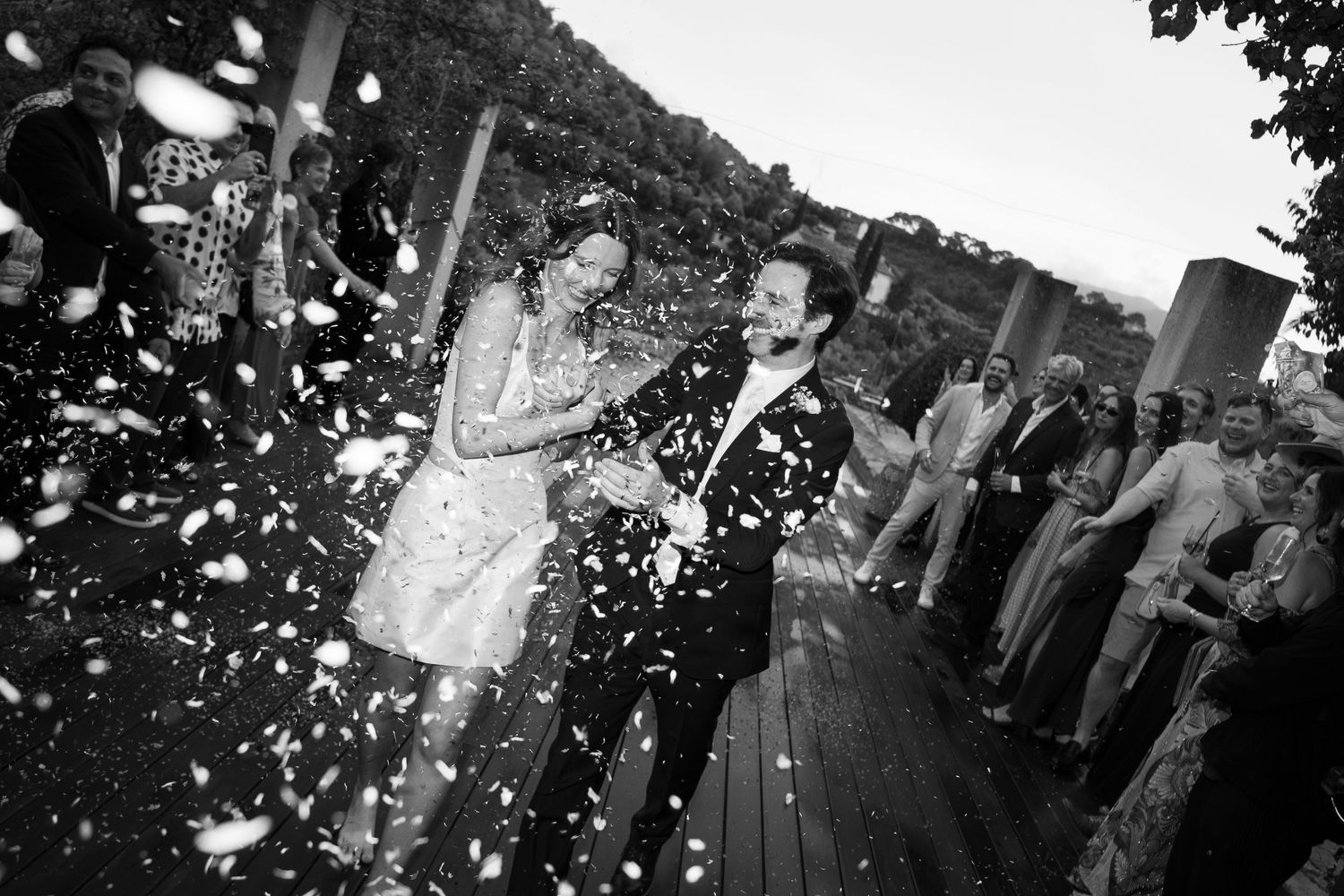 Black and white wedding photo of a bride and groom laughing as guests toss confetti on a wooden deck outdoors.