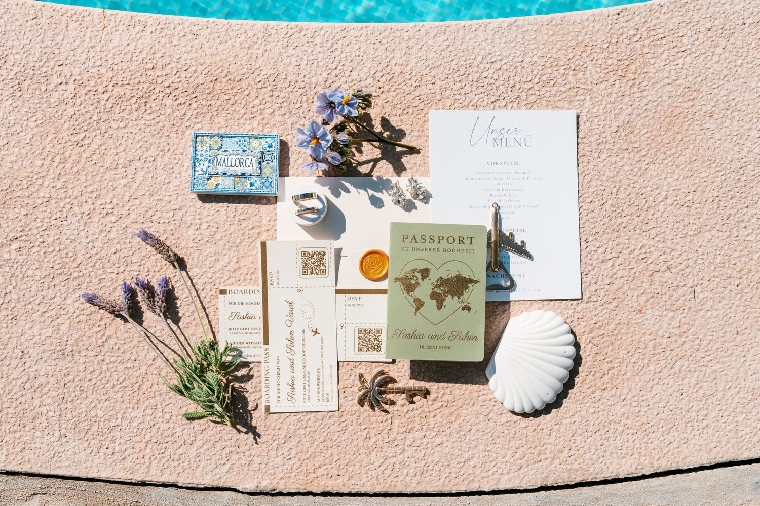 Travel-themed wedding invitation suite with passport design, lavender, and seashell displayed by a pool in Mallorca.