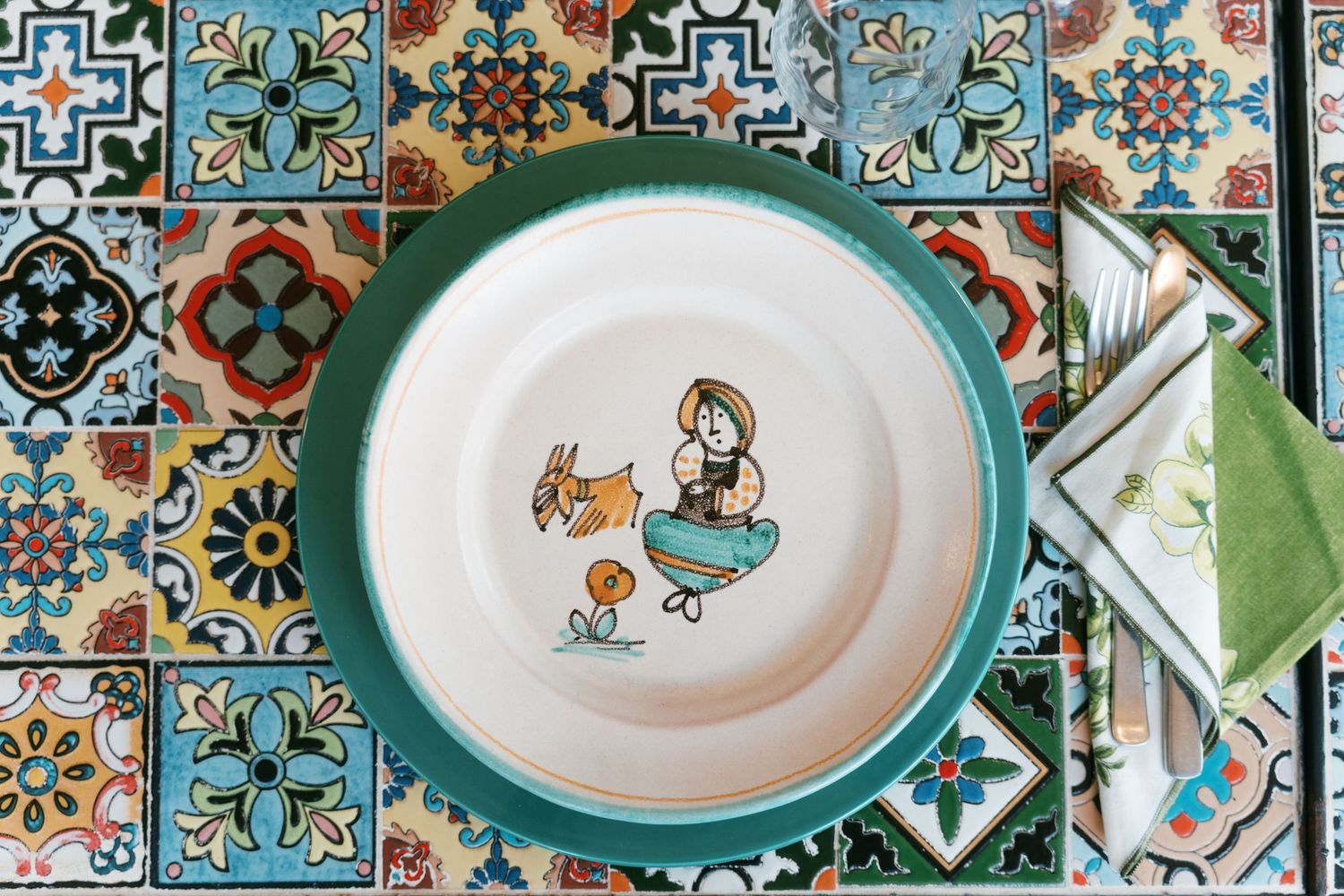 Colorful hand-painted ceramic plate with folk art design on a mosaic tile table with a folded napkin. Tuscany wedding photographer, filmmake