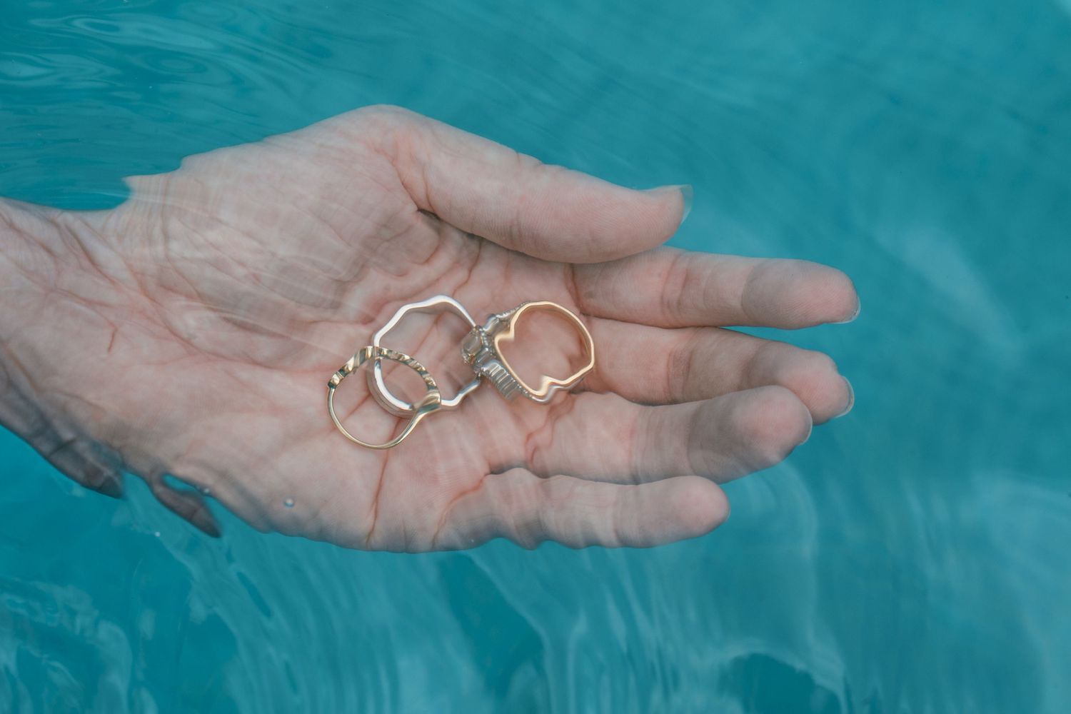 A hand holding delicate gold and rose gold heart-shaped rings over a turquoise swimming pool. Mallorca wedding photographer, filmmaker