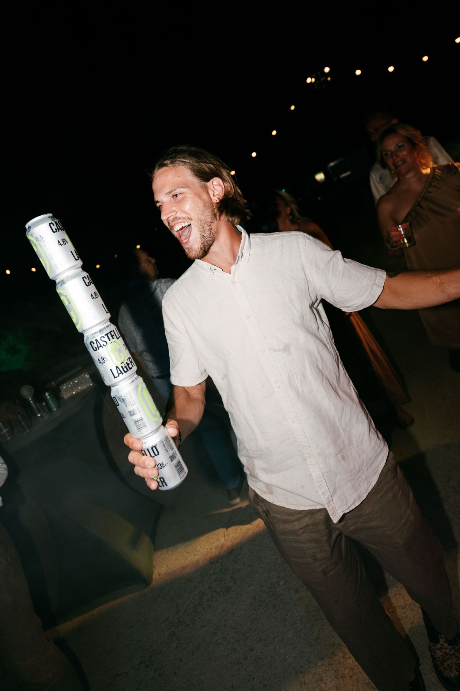 Man laughing while balancing stacked Castle Lager beer cans at an outdoor night party.