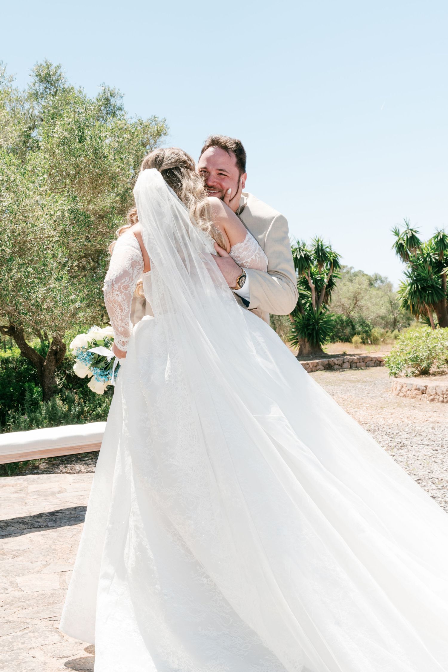 Groom in beige suit embracing bride in white lace gown and veil, holding bouquet, outdoors on sunny wedding day in Mallorca.