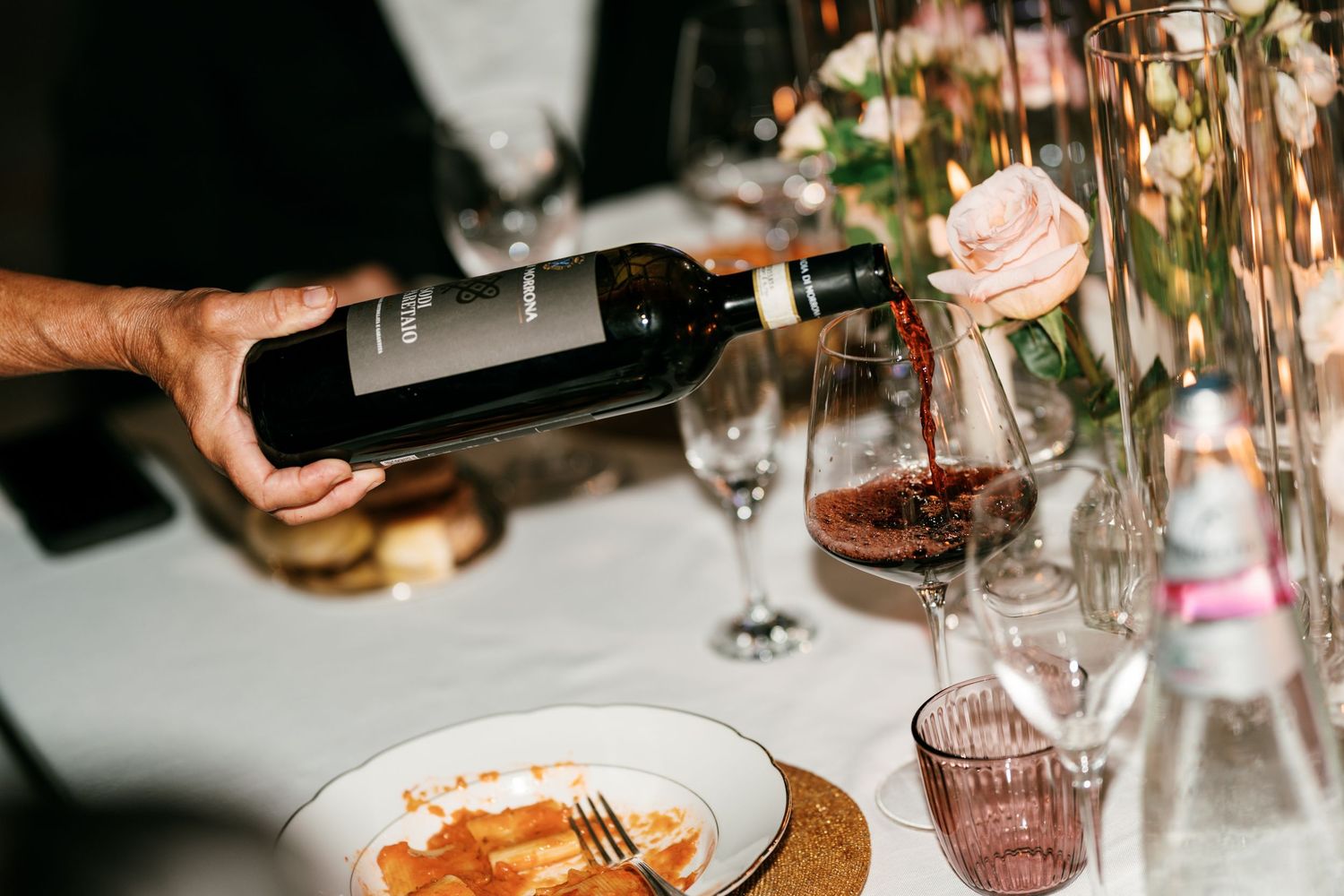 Hand pouring red wine from a bottle into a glass at a wedding dinner table with a pink rose and plate of food nearby.