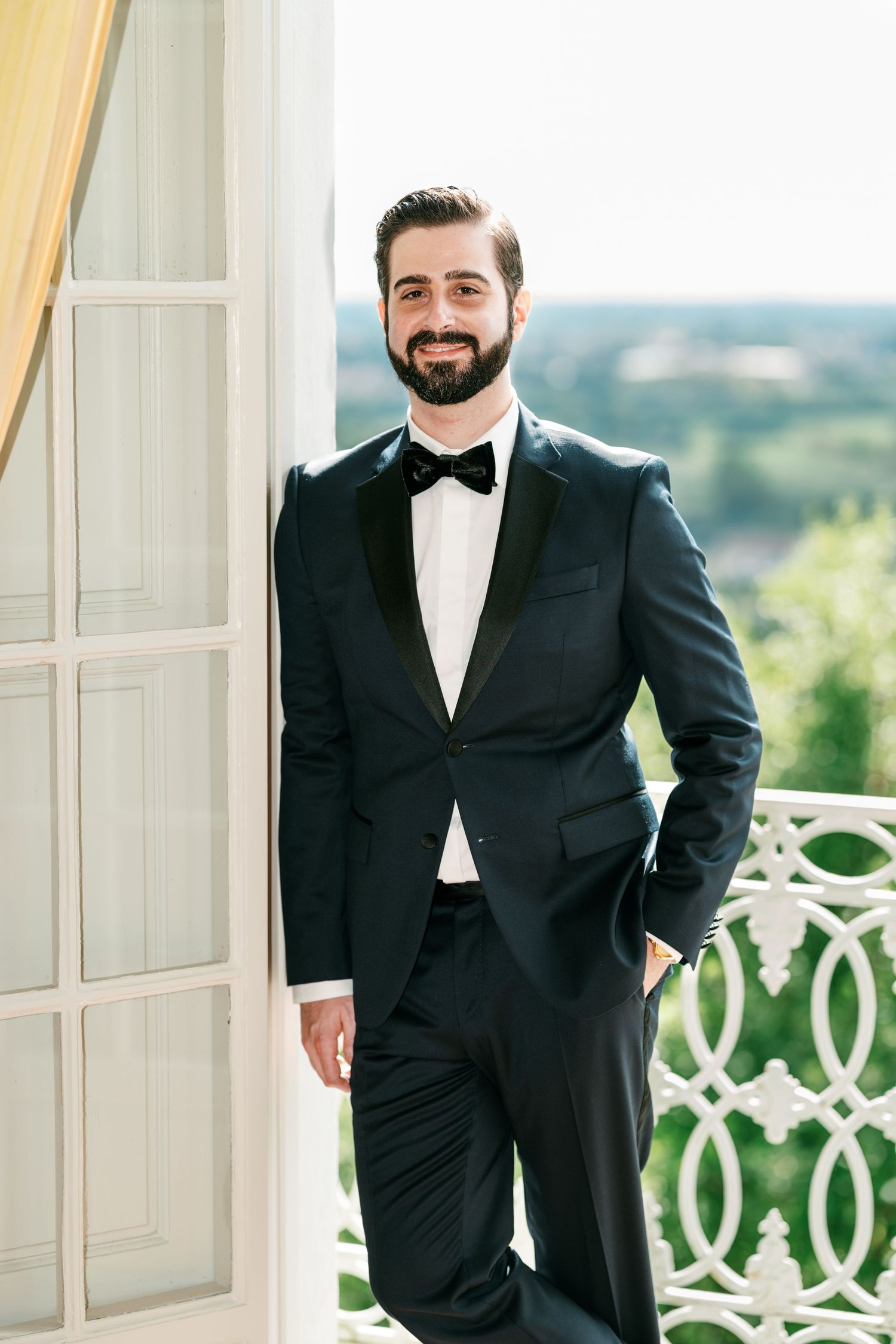 Smiling bearded man in a black tuxedo and bow tie leans against a white door beside an ornate balcony railing with a scenic view.