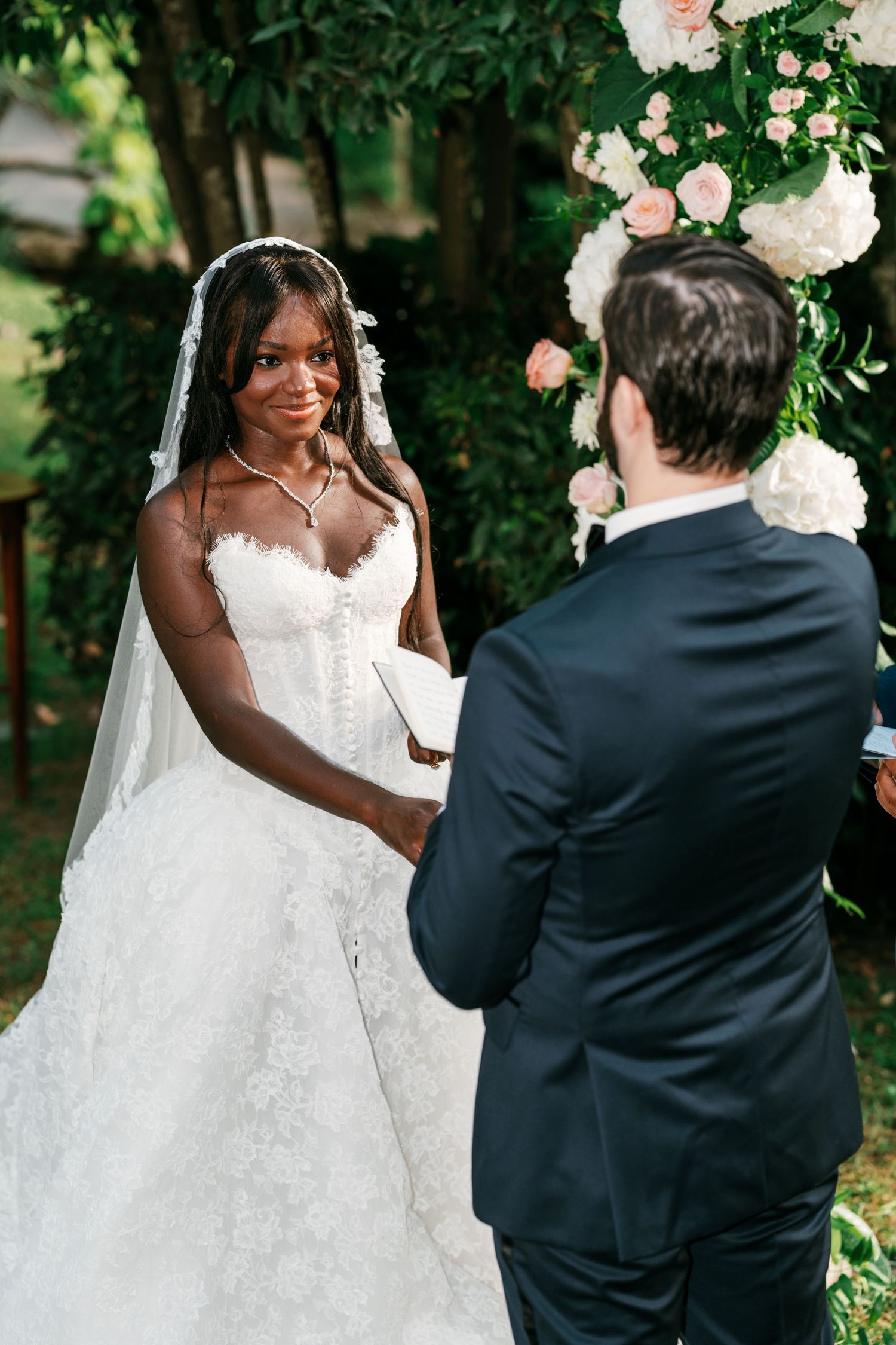 Smiling bride in a strapless lace gown and veil holds hands with the groom in a navy suit during an outdoor garden wedding ceremony.