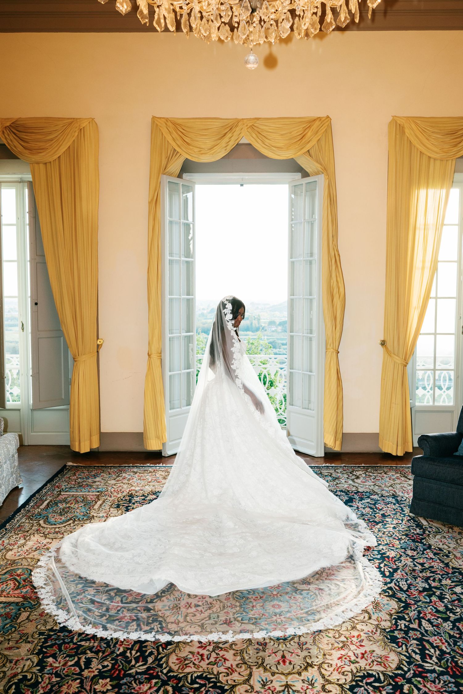 Bride in a lace wedding gown with a long cathedral veil and train stands before open French doors in an elegant room with yellow drapes.