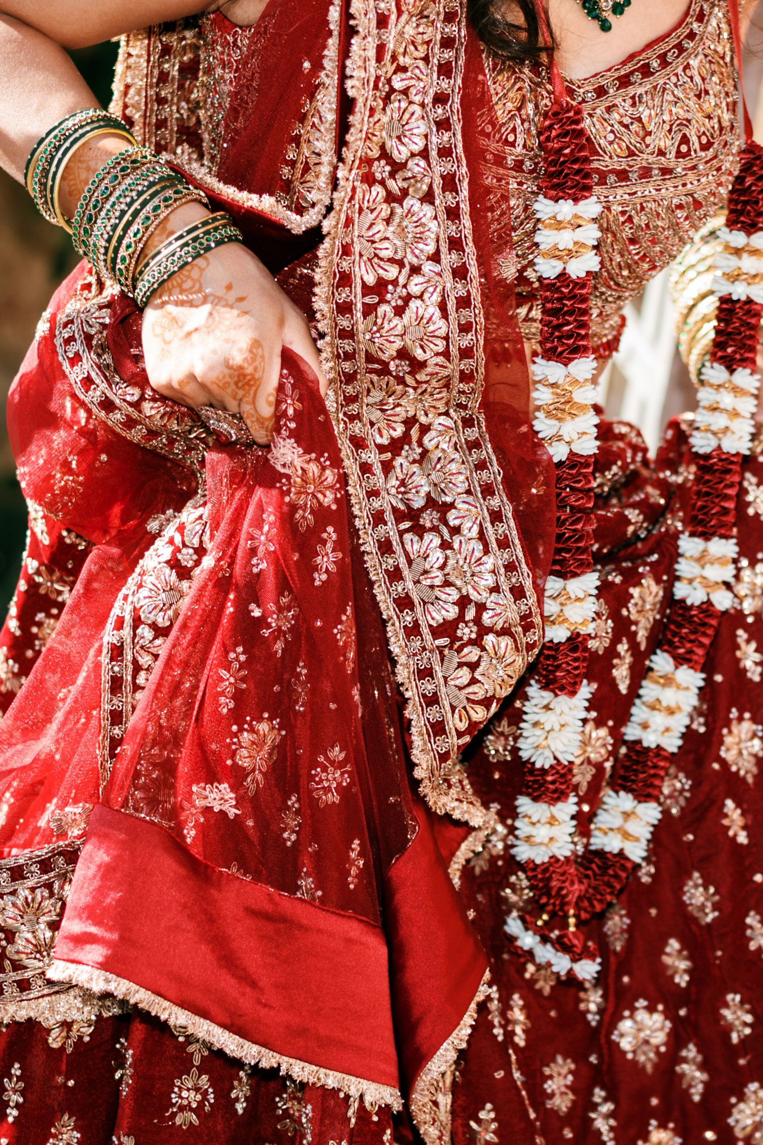 Bride in a red Indian wedding lehenga with gold embroidery, stacked bangles, henna-decorated hands and a white and red floral garland.