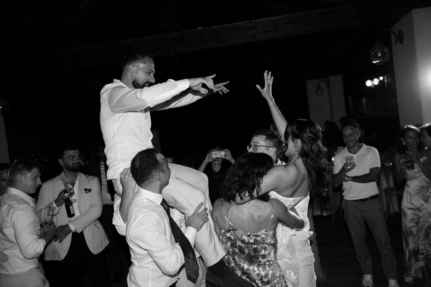 Black and white photo of wedding guests cheering and dancing as a man is lifted into the air on a chair at a lively reception.