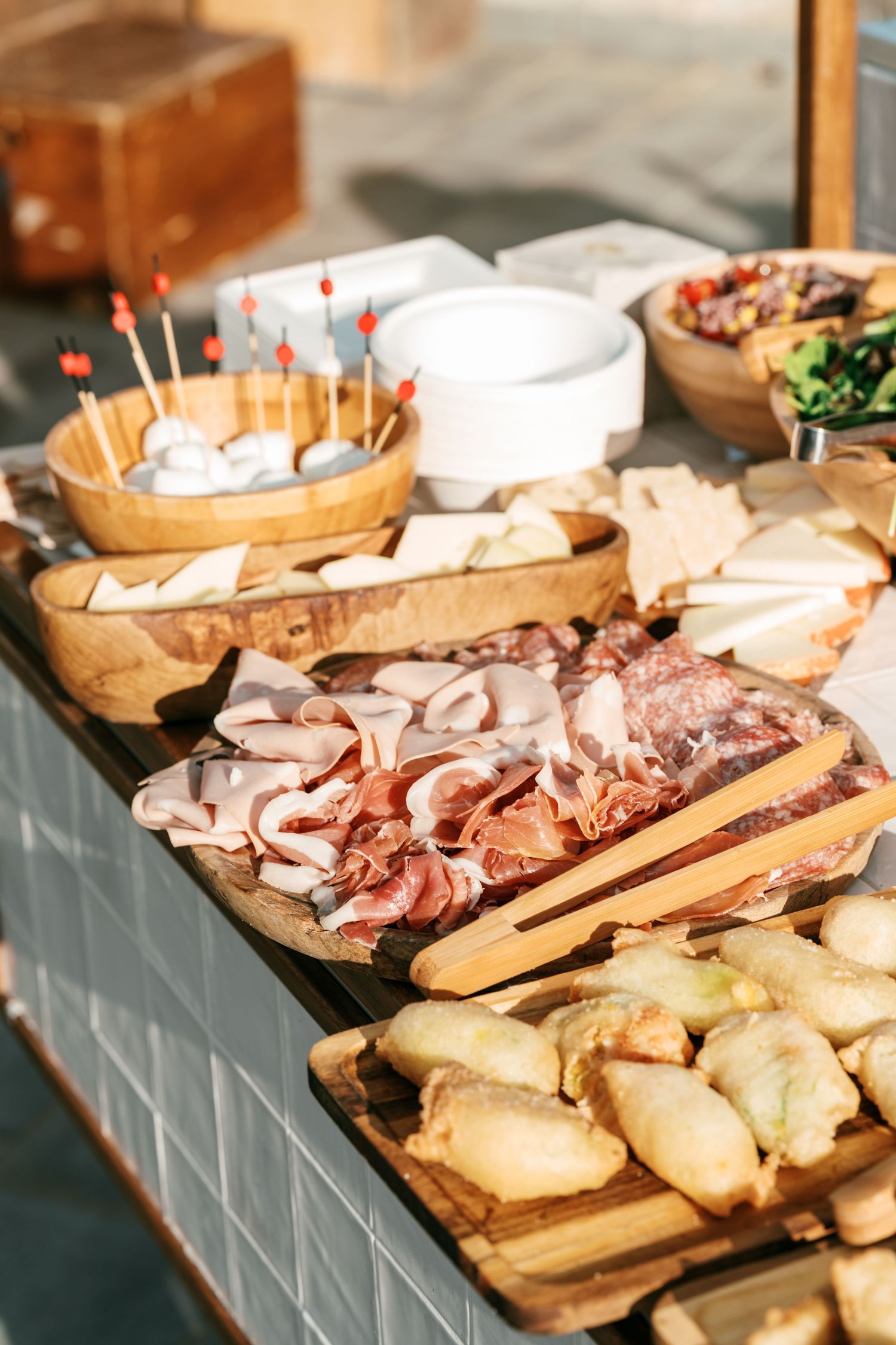 Rustic antipasto buffet with sliced cured meats, assorted cheeses, fried pastries, and appetizer skewers on wooden boards and serving trays.