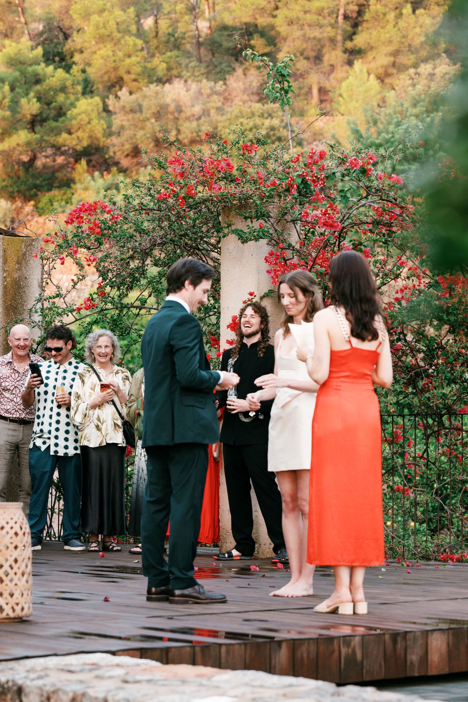 Bride and groom exchange vows on a wooden deck as guests watch, framed by red bougainvillea and wooded hills.