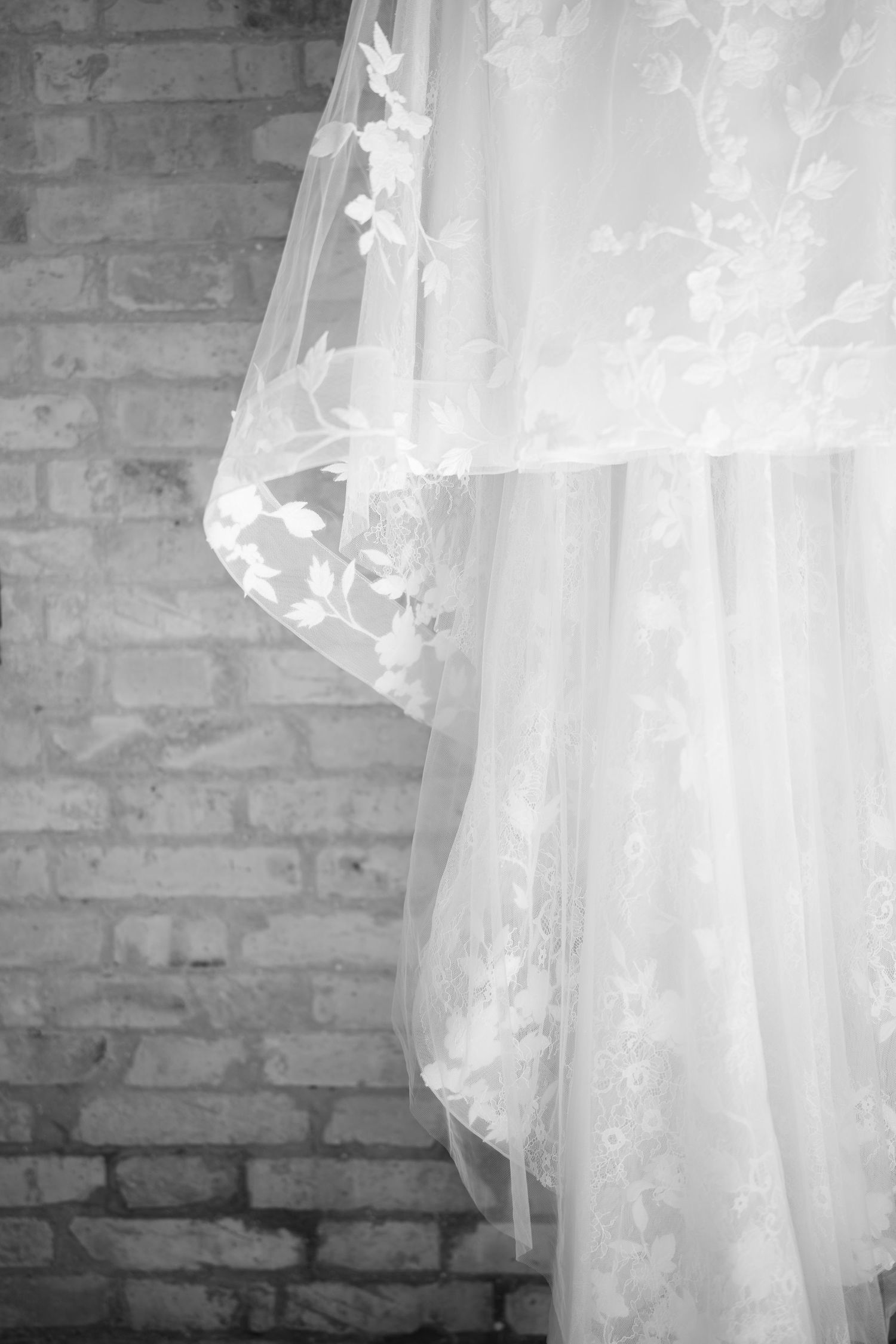 Black and white photo of a delicate lace wedding dress with floral veil hanging against a brick wall.
