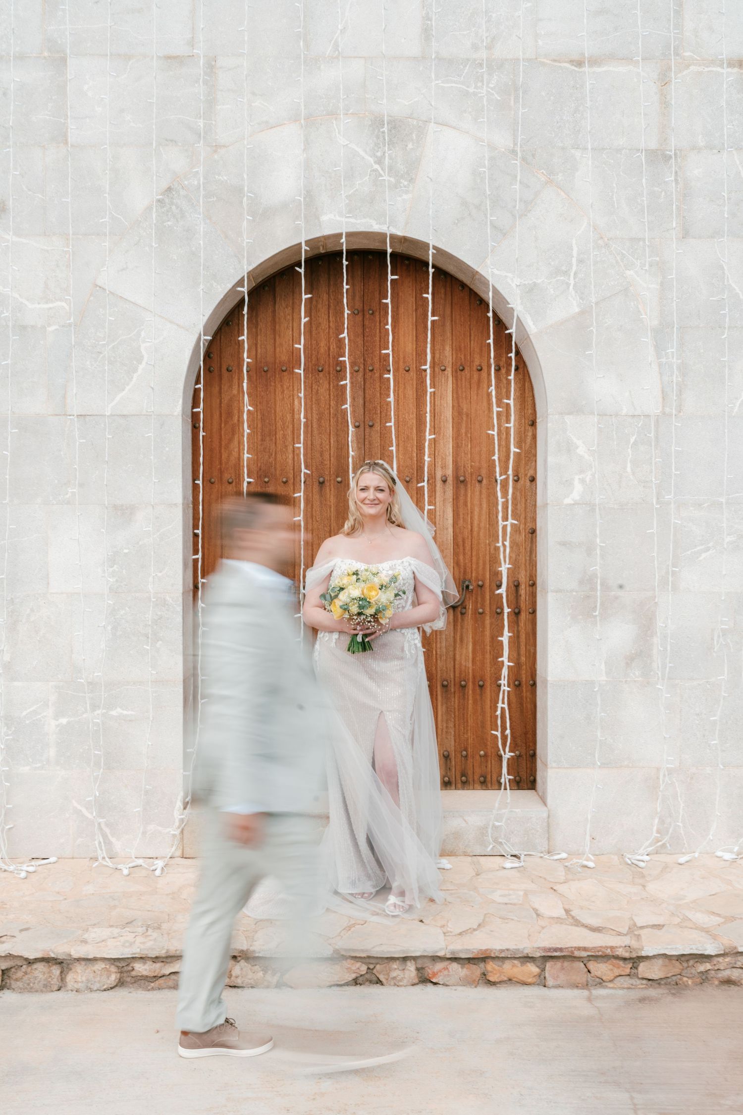 Bride in sheer gown holds yellow bouquet by wooden arched door as groom walks past in light suit.