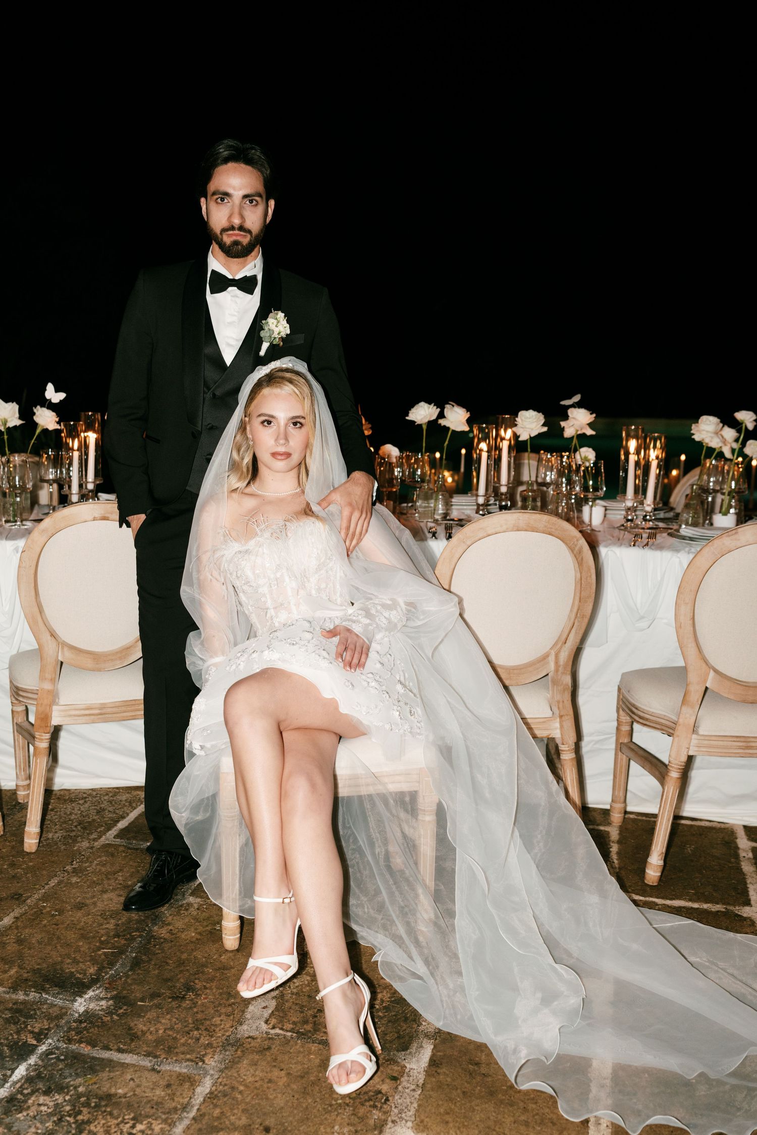 Elegant bride in lace mini dress and veil sits beside groom in black tuxedo at candlelit wedding reception table.