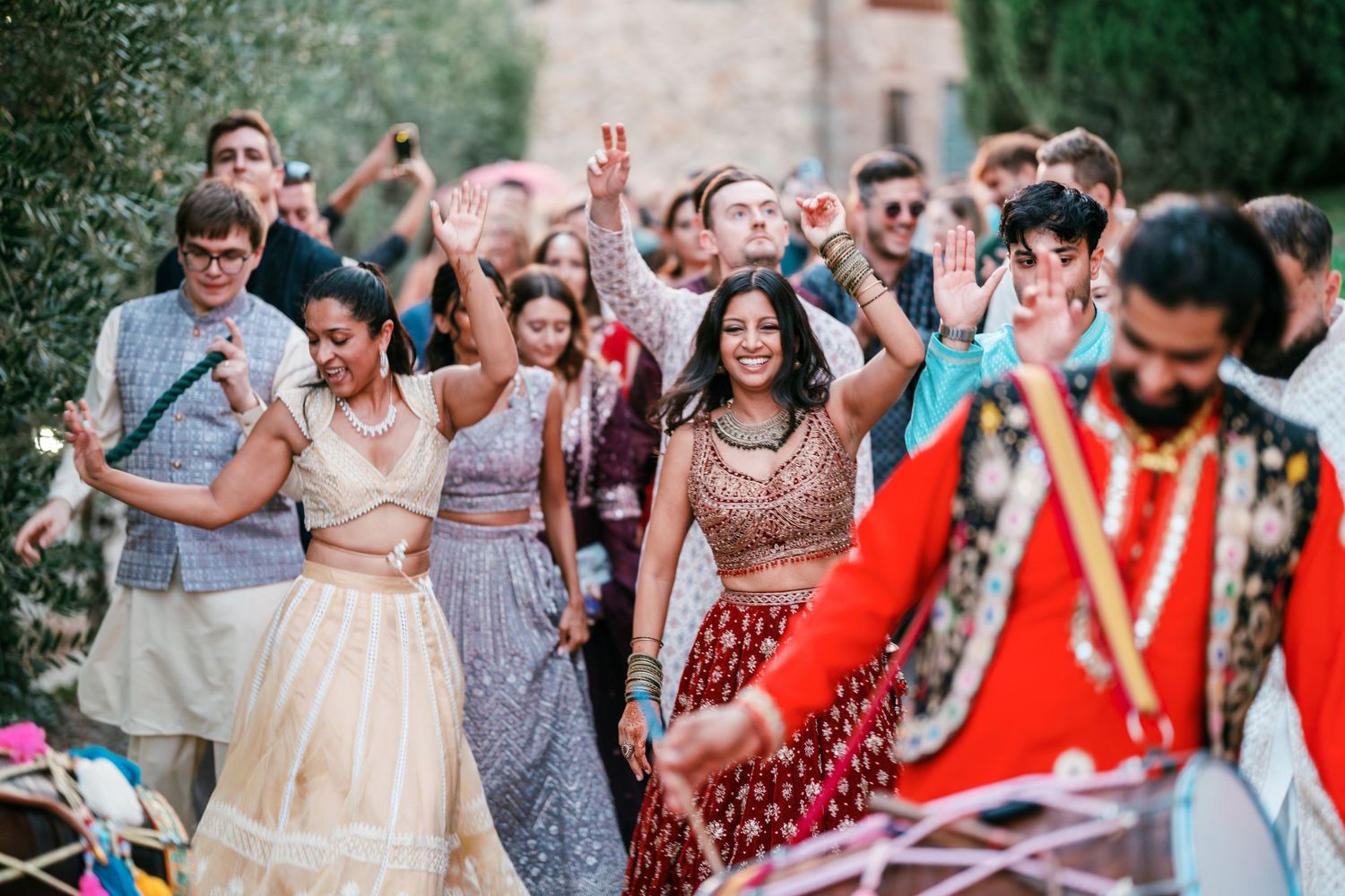 Guests in colorful traditional Indian attire clap and dance joyfully outdoors at a wedding celebration, with a drummer in the foreground.