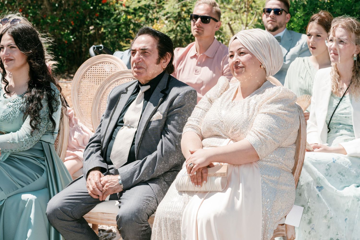 Guests in formal attire smile during an outdoor wedding ceremony, seated in ornate chairs surrounded by greenery in Mallorca.