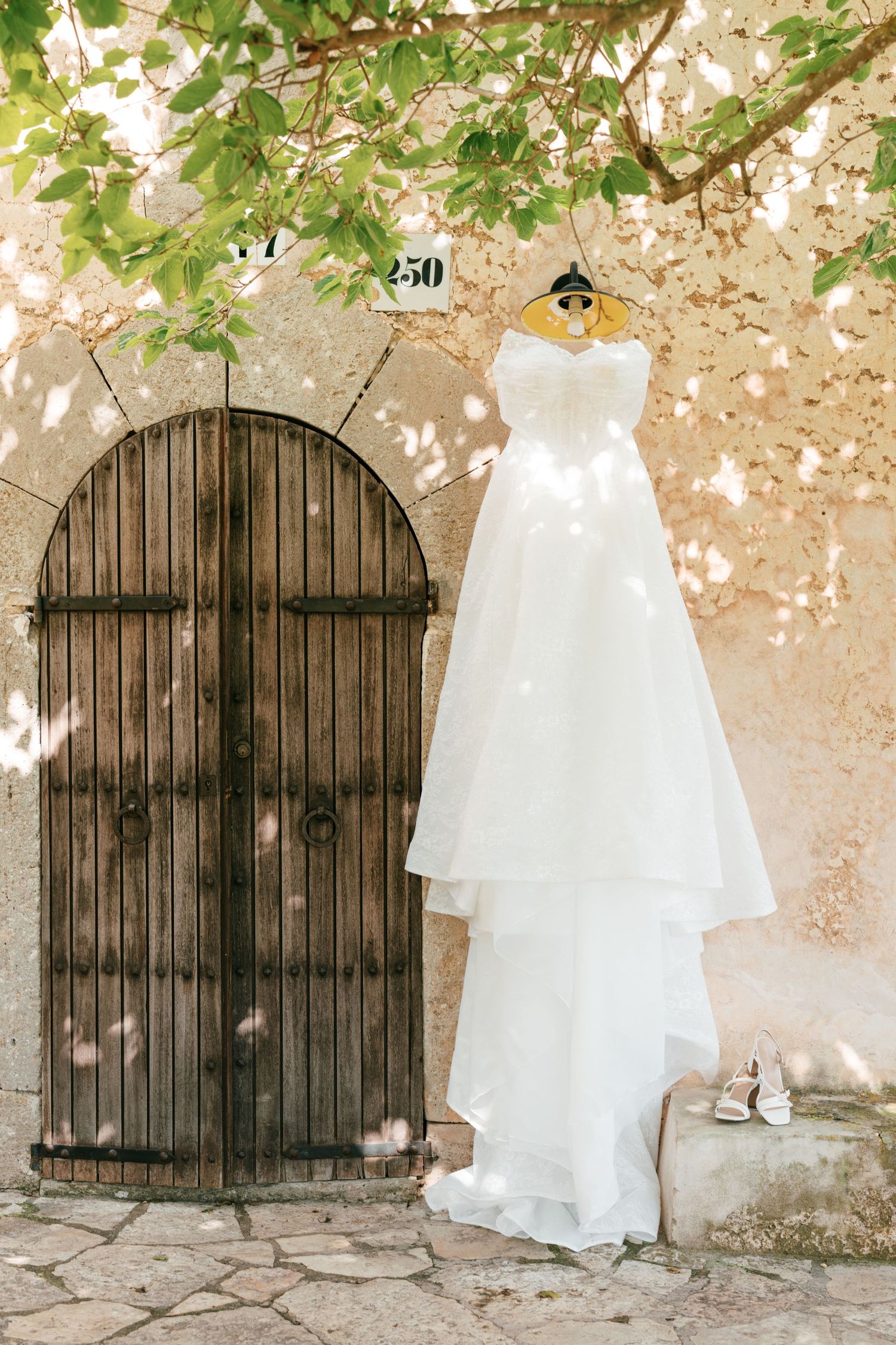 White strapless wedding dress hanging on rustic stone wall beside arched wooden door, with heels below in Mallorca.
